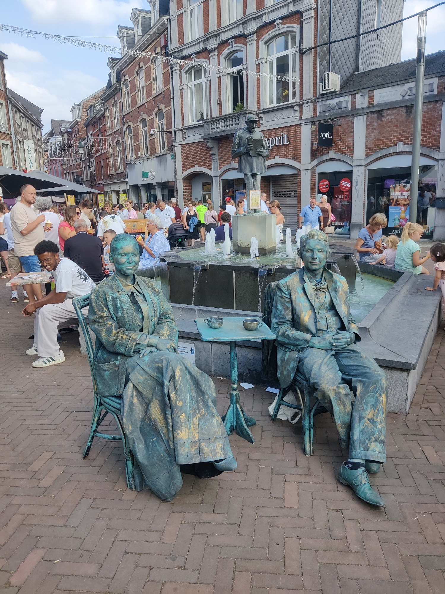 The image shows a street scene in Marche-en-Famenne, Belgium, featuring two human statues painted to resemble bronze figures. The statues are seated in chairs, with one to the left appearing to be a woman in a dress and jacket, and the other to the right a man in a suit and bowtie. They are positioned in front of a fountain. Numerous people are visible in the background, walking and gathered around the fountain, suggesting a public square or pedestrian zone. Buildings with red brick facades and storefronts line the street. The ground is paved with brickwork. The overall atmosphere appears to be a lively, outdoor public gathering.