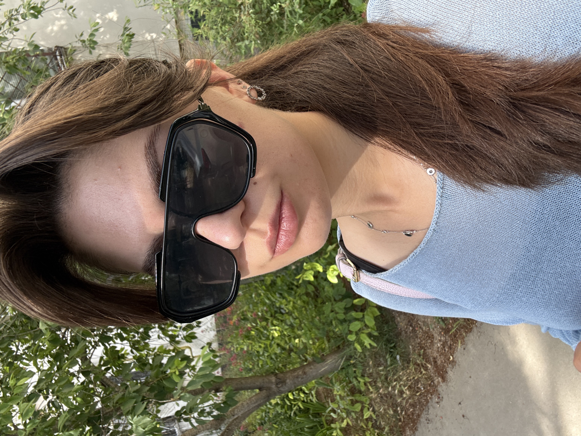 A woman is taking a selfie outdoors. She is wearing sunglasses, a light blue sweater, and a necklace. Her hair is brown and pulled back in a ponytail. There is a tree and some bushes in the background.