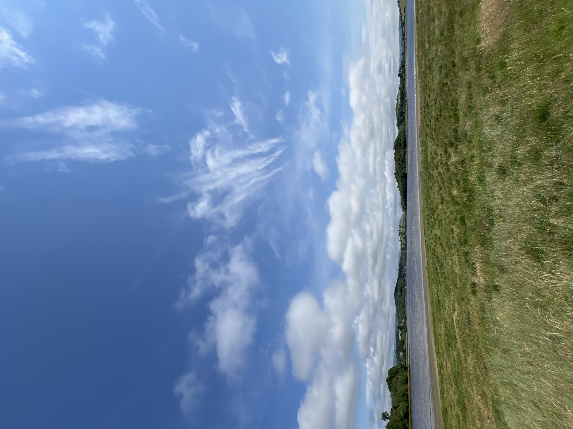 The image captures a scenic view of a body of water, likely a bay or estuary, bordered by lush green grass in the foreground. In the distance, rolling hills covered in trees and vegetation create a picturesque backdrop. The sky is a vibrant blue, dotted with a mix of fluffy white clouds and wispy cirrus formations, suggesting fair weather. The water is calm, reflecting the sky and the distant landscape. The overall impression is one of a serene and natural environment, possibly in a rural or semi-rural setting. The location is identified as Mountcharles, Ireland.