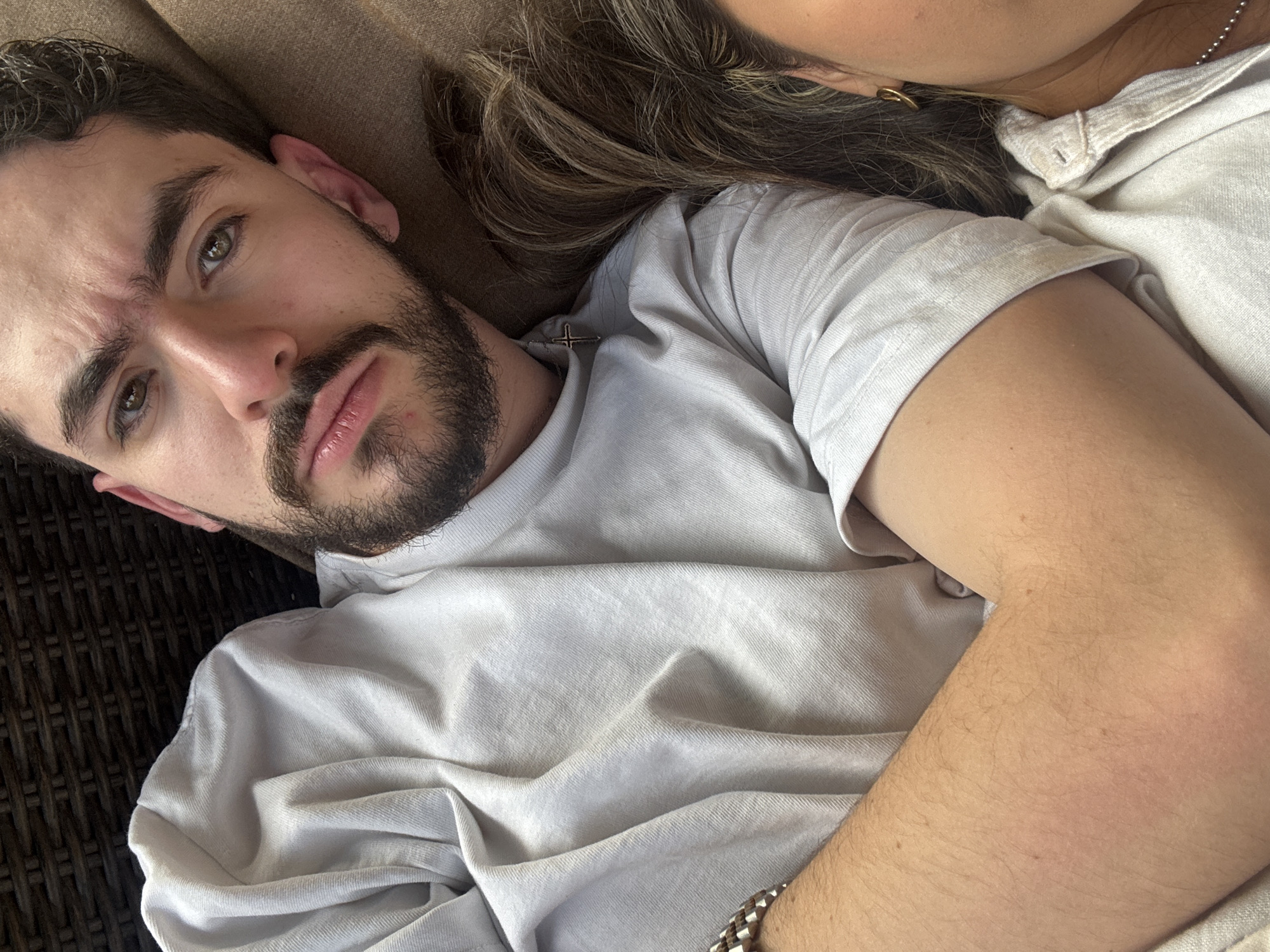 A close-up, eye-level shot depicts a man with dark hair and a beard, lying on what appears to be wicker furniture. The man is wearing a light-colored t-shirt. Beside him, a woman is visible, with part of her face and shoulder showing, also wearing a light-colored shirt. The location and city are unknown.