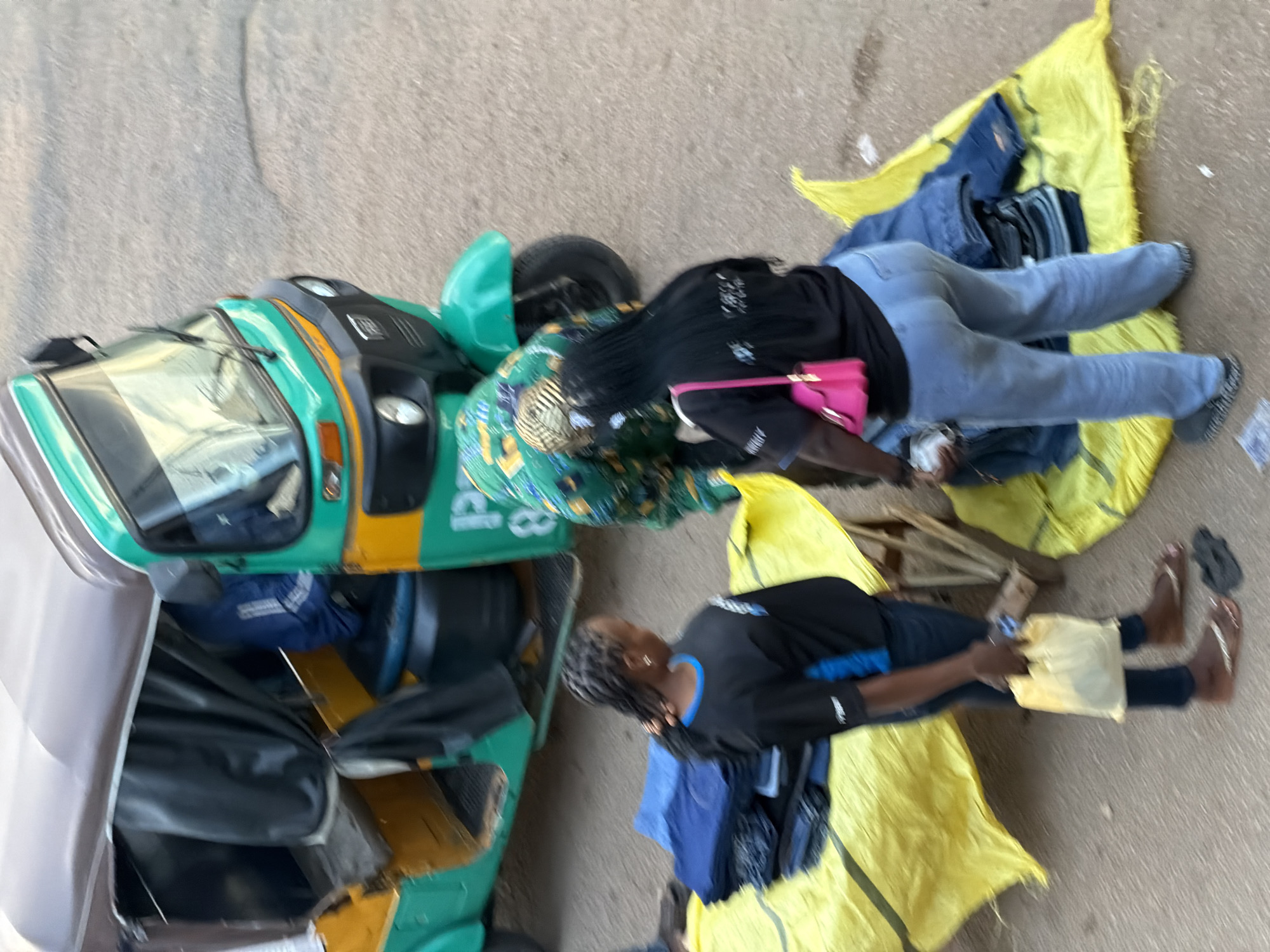 In Bukuru, Nigeria, a scene unfolds on a dusty, uneven surface, likely a roadside or market area. Two individuals, both women, are engaged in what appears to be commerce. One woman, wearing a green and yellow patterned top and black pants, is seated behind a three-wheeled vehicle, possibly a tuk-tuk, which is primarily green and yellow. The vehicle's front section is visible, with its windshield and headlights. A bright pink purse is slung across her shoulder. Beside her, a large, bright yellow tarp is spread out, laden with folded blue jeans and other clothing items.
The second woman, dressed in a black t-shirt with a blue collar and dark pants, is sitting on another yellow tarp, which also displays clothing. She is holding a small, light-colored object, perhaps a receipt or a small item for sale. Her posture suggests she might be interacting with a customer or tending to her wares. The overall impression is one of a bustling, informal marketplace. The lighting suggests it is daytime, with no clear indications of weather conditions beyond the dry ground. The focus of the image is on the women and their merchandise, highlighting a moment of daily economic activity.