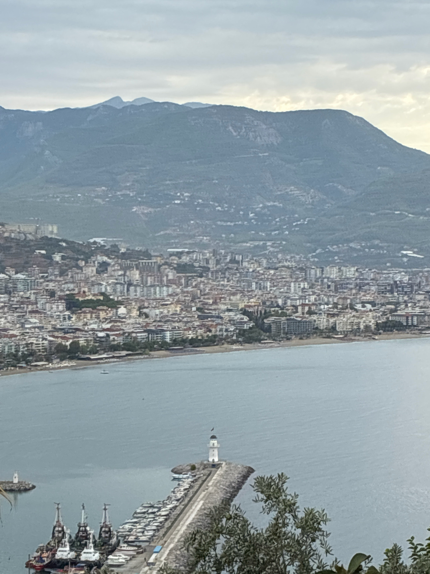 This is a long-range, high-angle, eye-level, medium shot of the Alanya coastline in Turkey. The scene is overcast. In the foreground, on the right, is a dense cluster of green foliage. To the left of the foliage is a body of water with several boats docked on a pier. The pier is lined with yachts and is topped by a white lighthouse with a black roof. Further left, in the water, is another, smaller lighthouse.
In the mid-ground, there is a coastline with a sandy beach, dotted with umbrellas and lounge chairs. Beyond the beach is a densely packed city with numerous buildings of varying heights and colors. A large hill or mountain range dominates the background, with a hazy, overcast sky above. The city appears to stretch along the coastline and up the slopes of the hills. Several cruise ships or large vessels can be seen docked in the harbor area.