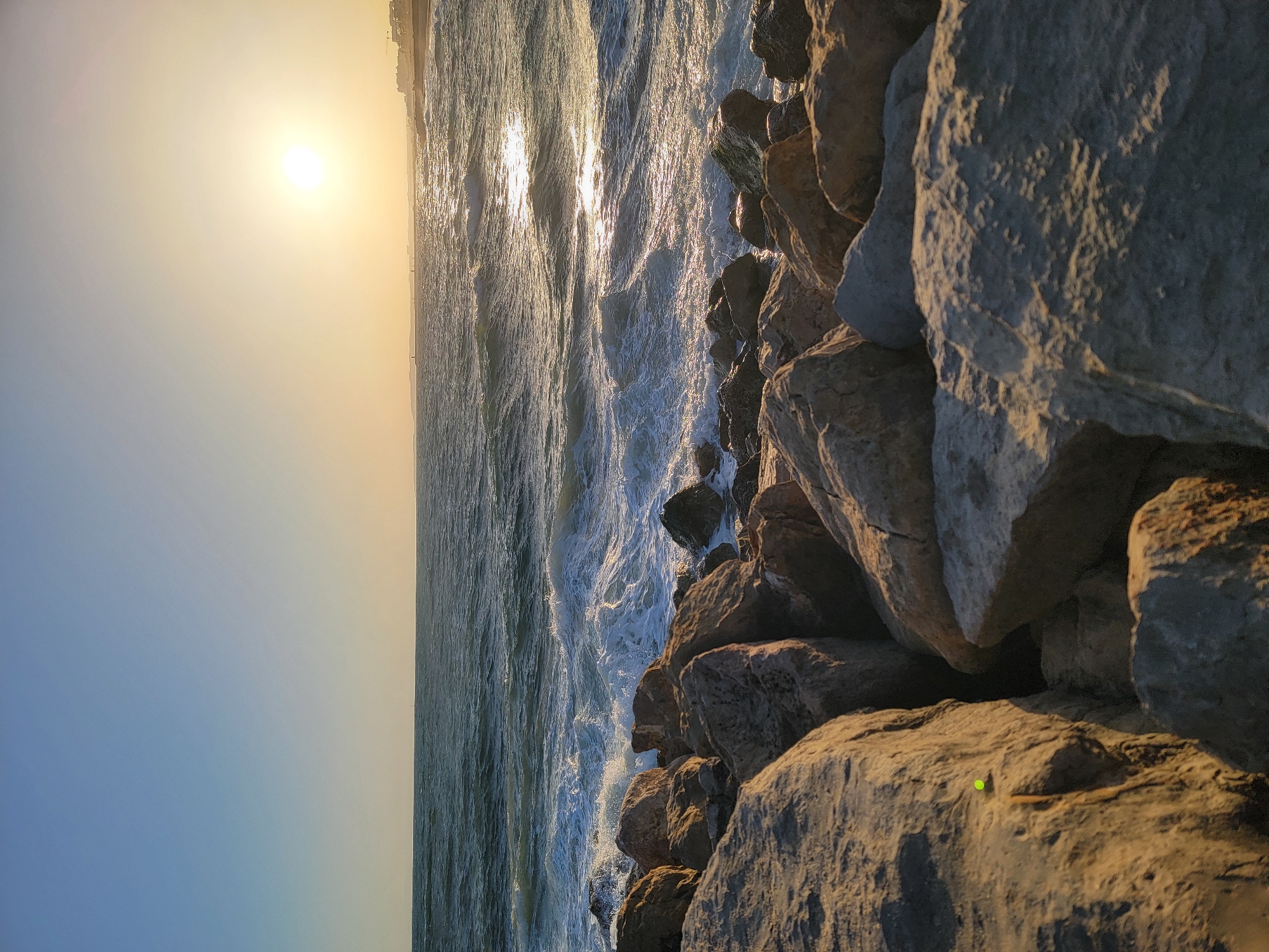 This is a horizontal, low-angle, close-up, wide shot of a rocky coastline at sunset. The sun is low on the horizon, casting a warm, golden light across the scene. The sky transitions from a pale blue at the bottom of the frame to a hazy yellow and orange as it approaches the sun. The sun itself is a bright, orb-like sphere, its light reflecting off the choppy water of the ocean.
The foreground is dominated by large, rough-textured rocks, characteristic of a breakwater or natural rock formation. These rocks are illuminated by the setting sun, with warm highlights and deep shadows that emphasize their ruggedness. In the middle ground, the ocean is a vibrant blue-green, with waves crashing against the shore, creating white foam and spray. The sunlight glints off the surface of the water, creating streaks of light. In the distance, there's a faint line of buildings visible along the horizon, suggesting an urban or coastal settlement.
The overall mood is peaceful and serene, with the warm colors of the sunset evoking a sense of calm and tranquility. The time of day is clearly sunset due to the low angle of the sun and the warm, golden light. The weather appears clear, with no clouds obscuring the sun. The presence of the ocean and the rocky shore strongly suggests a coastal location. While there are no visible people or animals, the faint line of buildings hints at human presence in the vicinity. No text is visible in the image.
