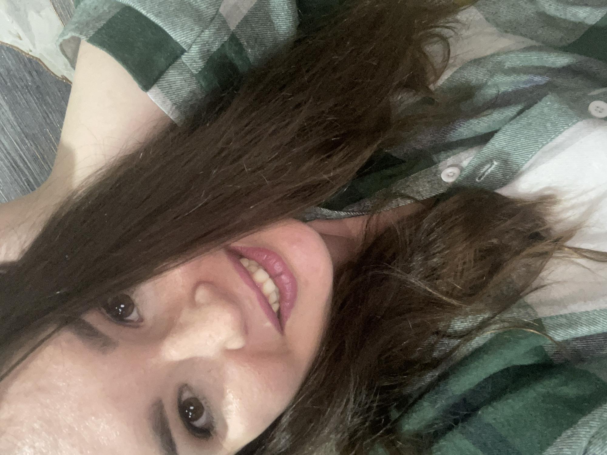 A woman with long, dark brown hair and brown eyes is angled and smiling towards the camera. She is wearing a green and white plaid shirt. The background appears to be a light-colored surface. There is no evidence to suggest this image was taken in the city of Chaikovsky, Russia.