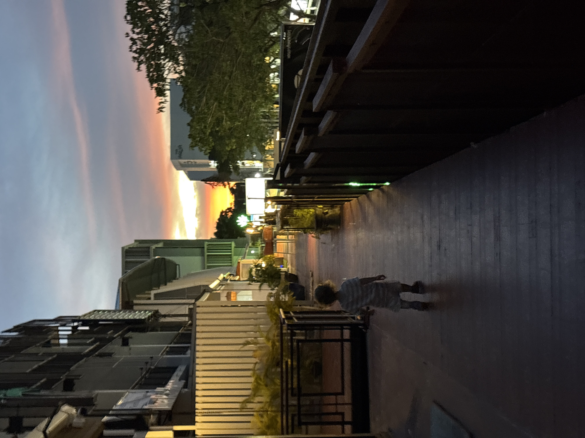 A child walks on a wooden walkway in Choeng Thale, Thailand, at dusk. The sky displays a gradient of sunset colors, from orange and yellow near the horizon to pink and blue higher up. Buildings with balconies and windows line the left side of the walkway, while a dark wooden fence borders the right. Various potted plants are visible along the walkway, and some lights are illuminated, indicating the early evening. The overall atmosphere is tranquil, with the fading light creating long shadows.