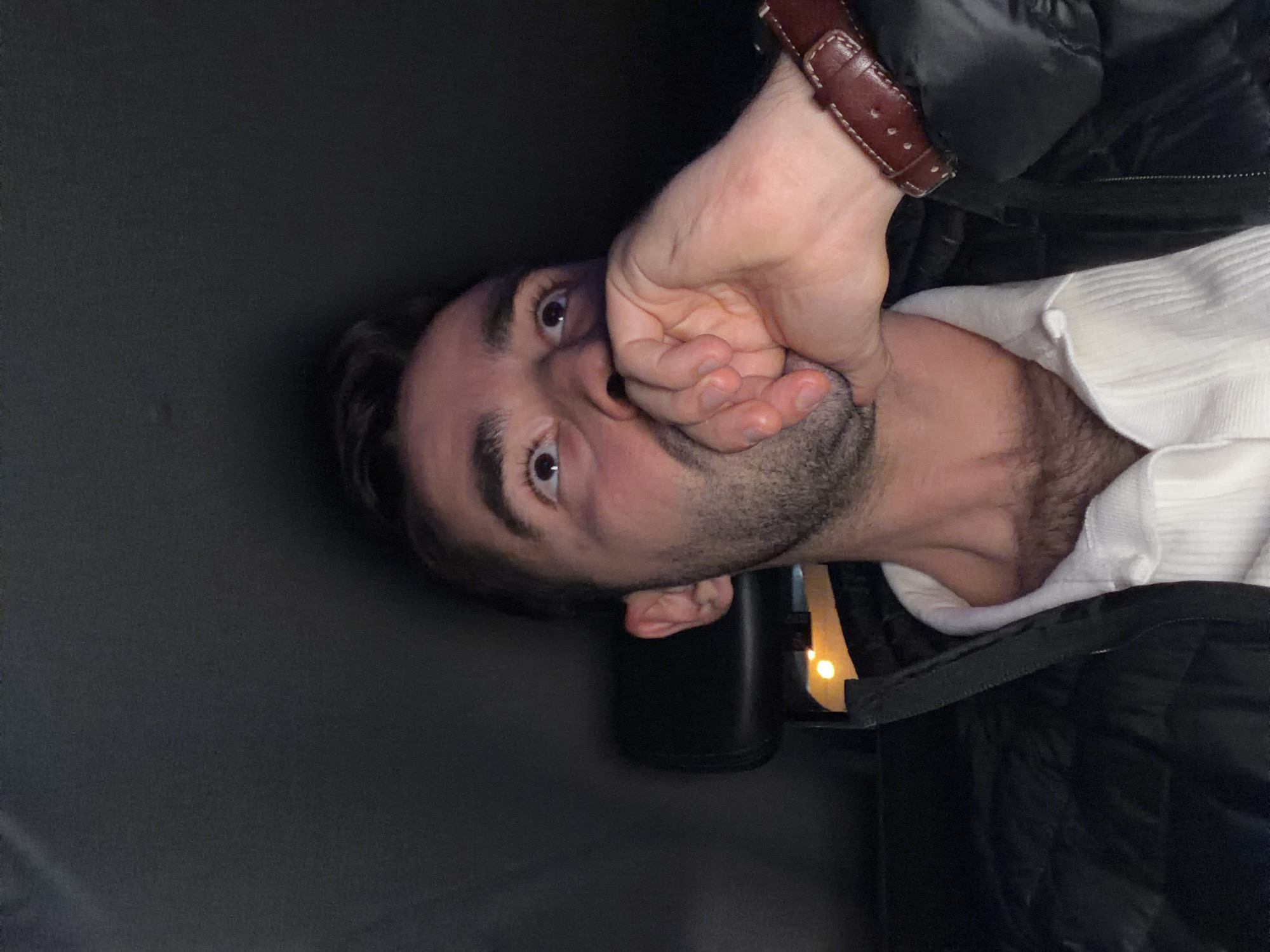 A man with dark hair and eyes is shown close-up, partially covering his mouth with his hand. He is wearing a white sweater under a black puffer jacket. A brown leather watch is visible on his wrist. In the background, a black container with a glimpse of light can be seen. The image appears to be taken in a dark environment. The location cannot be determined solely from the provided image. There is no indication the location is in Moscow, Russia.
