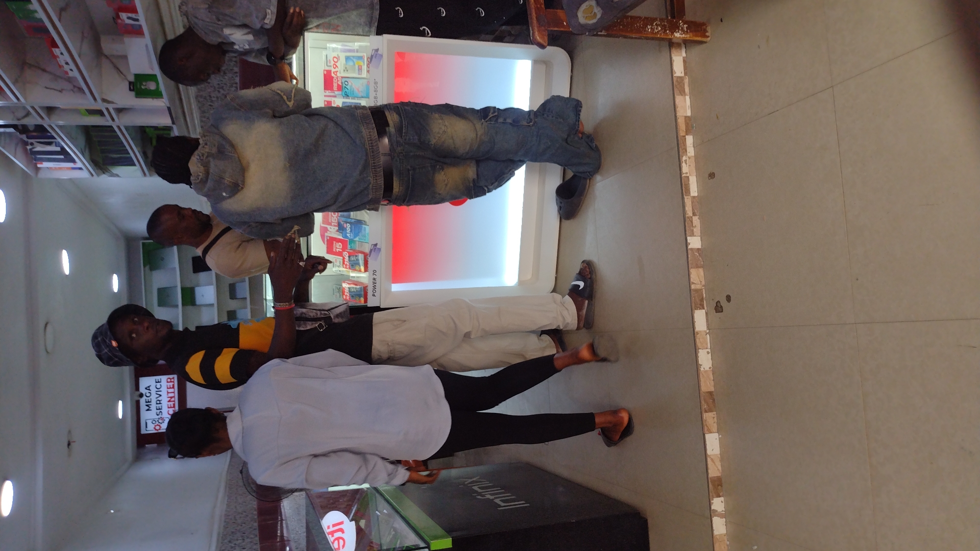 The photograph was taken in what appears to be a retail store, possibly a mobile phone shop, judging by the "Infinix" branding and the "MEGA SERVICE CENTER" sign visible in the background. Four individuals are present. One wears a light-colored shirt and black leggings, standing near a display case with the Infinix logo. Another person is wearing a black shirt with yellow stripes and is closer to the MEGA SERVICE CENTER sign, also facing forward. Two more individuals are near a lighted display case showcasing mobile phones, with details like "RED 490," "POWER 70," and "A100" visible. One wears a light cream-colored shirt and white trousers, and the other wears a denim jacket and jeans. The setting appears to be a tiled retail space with fluorescent lighting and possibly shelves of products in the background. The city of Jos, Nigeria, is mentioned, but there is no other information to confirm the location.