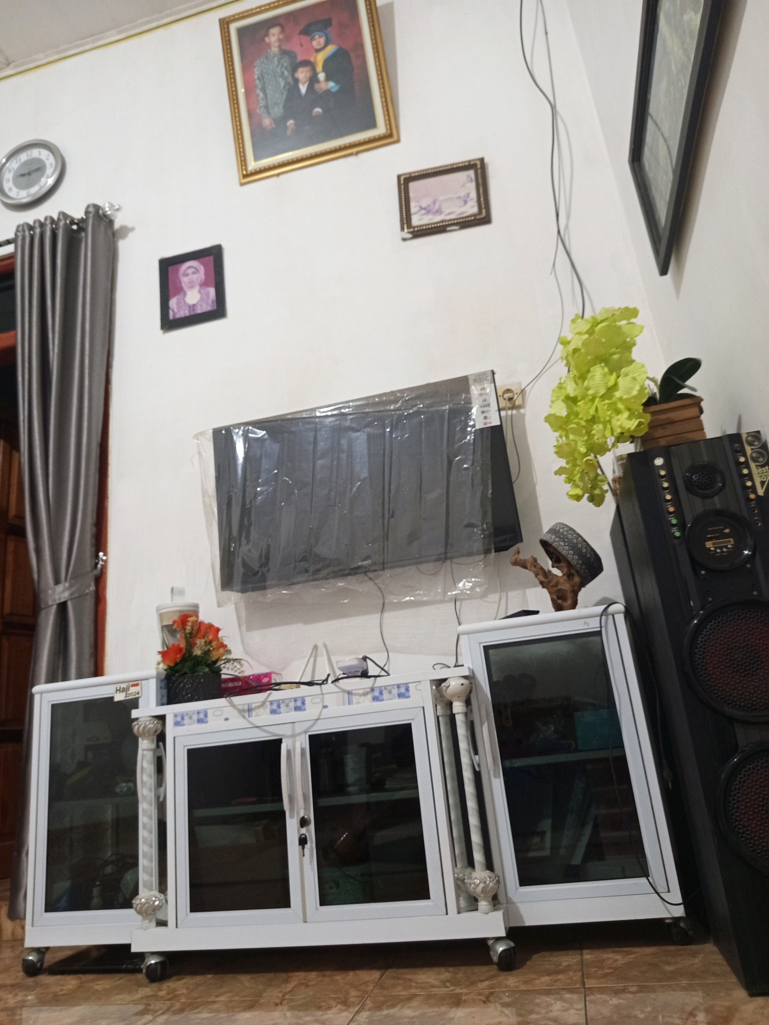 The image shows an interior scene with a white television stand with glass doors. A black speaker is positioned to the right of the television stand. On the wall above the television stand, there is a framed family photograph and two smaller framed pictures. A decorative plant with green leaves is placed to the right of the television. A round clock is visible on the left wall. A gray curtain hangs on the left side of the image. The floor appears to be tiled. The location is identified as Murni Jaya, Indonesia.