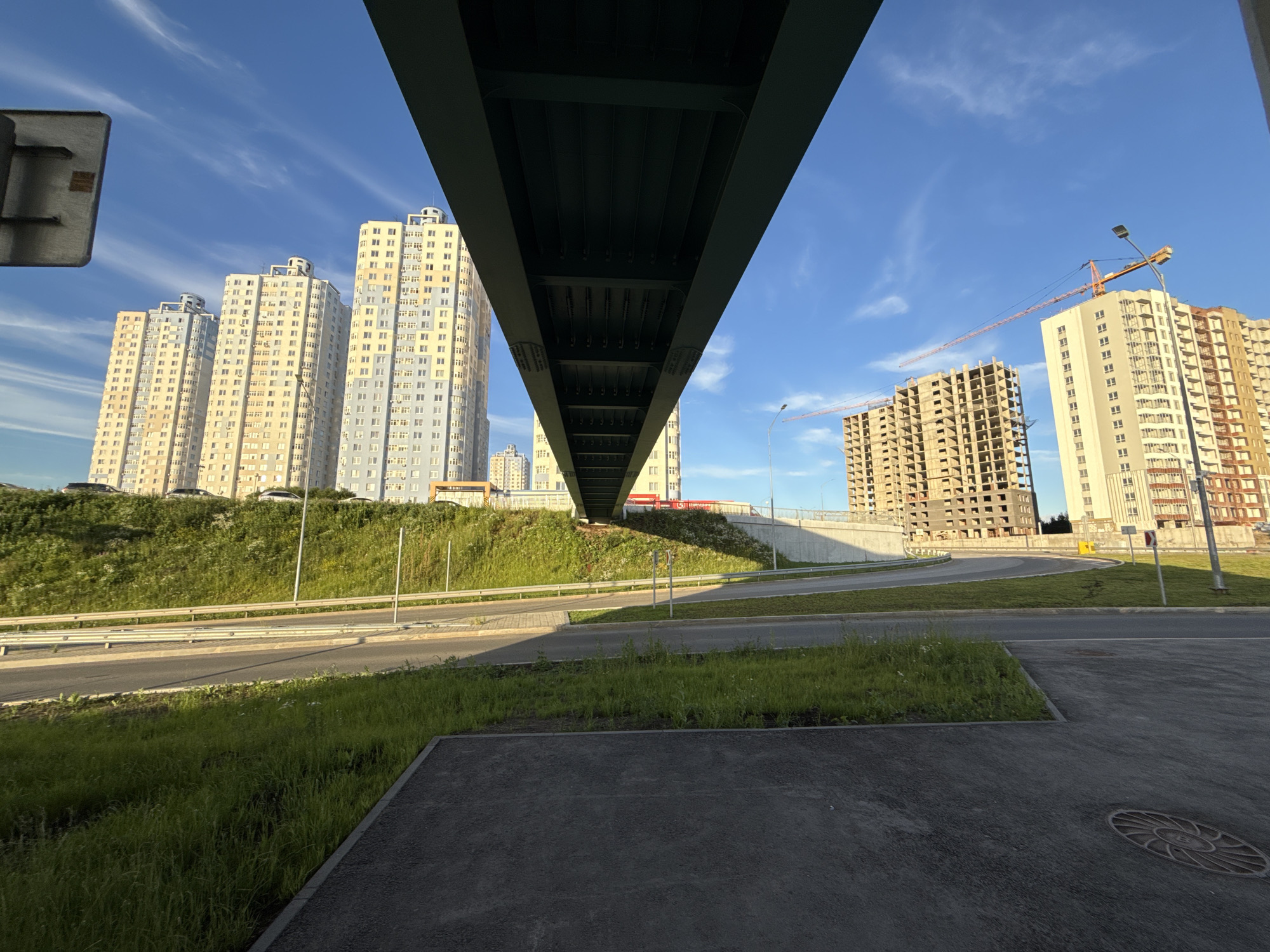 The media file depicts an urban construction scene under a clear blue sky with wispy white clouds. The perspective is low, looking up towards a large bridge structure that spans across the frame. Several tall, modern apartment buildings, some completed and others under construction, dominate the background. Two orange construction cranes are visible, one extended horizontally and the other positioned vertically, signifying ongoing development. The surrounding area includes a paved road, a grassy verge, and a sloping, overgrown embankment. The lighting suggests it is late afternoon or early evening, with the sun casting long shadows. There is no visible text in the media file. The scene conveys a sense of progress and urban expansion in a suburban or newly developed area.