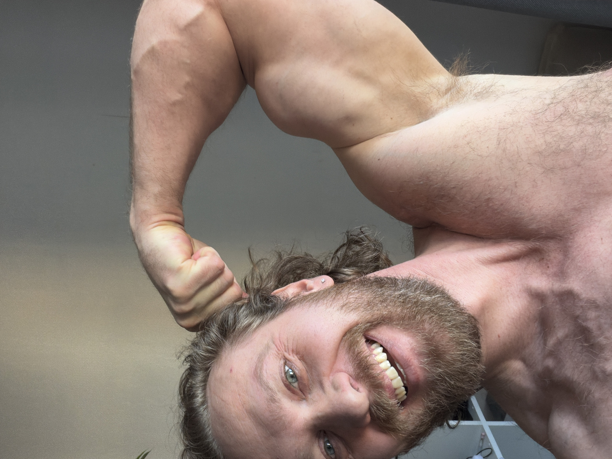 A shirtless man with a beard and fair skin is flexing his right arm, which is held above his head. His head is angled downwards, and he is smiling, exposing his teeth. He has green eyes and light brown hair. The background is a plain wall, and some white objects are visible behind him. There is no evidence to support that this image was taken in Saint Petersburg, Russia.