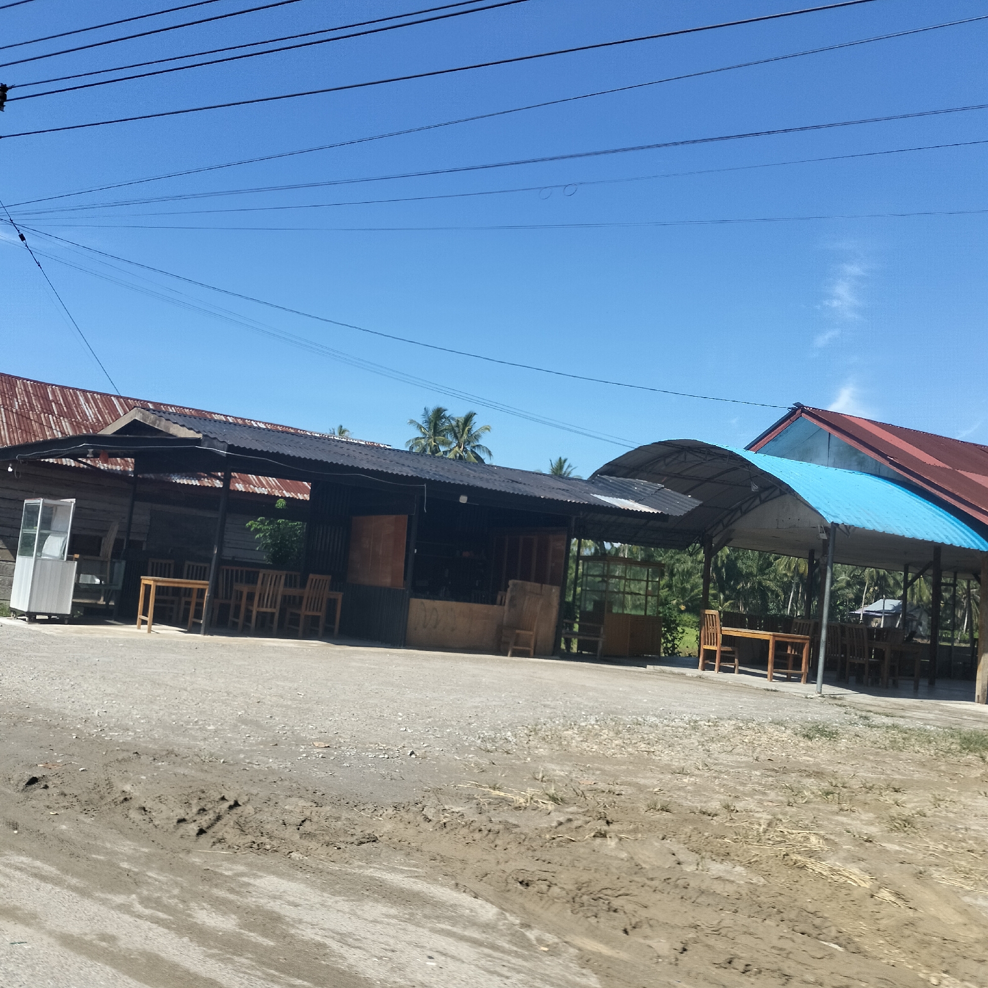 This image captures a bright, sunny daytime scene in Meunasahbeunot, Indonesia, featuring several open-air roadside eateries or cafes. The foreground consists of an unpaved surface, a mix of dirt, gravel, and some muddy areas with visible tire tracks.
To the left, a rustic building with wooden walls and a rusty reddish-brown corrugated metal roof is partially seen. Adjacent to it is an open-sided structure covered by a dark, possibly black, corrugated metal roof, supported by dark posts. Under this shelter, several light-colored wooden tables and chairs are arranged for dining. A white, glass-fronted display cabinet stands near the wooden building. Further right, another similar open-sided structure with a dark corrugated roof is visible, also equipped with wooden seating.
On the far right, a large, bright blue corrugated metal canopy extends over another seating area, featuring more wooden tables and chairs. Behind this blue canopy, a building with a red corrugated metal roof can be seen. The background is lush with numerous palm trees, indicative of a tropical environment.
The sky is a clear and vibrant blue with only a few faint wisps of clouds. Multiple black utility wires stretch across the upper portion of the frame. There are no people visible in the image, suggesting the establishments are currently unoccupied or closed. The scene presents a quiet, rural atmosphere under clear weather conditions. No visible text is discernible.