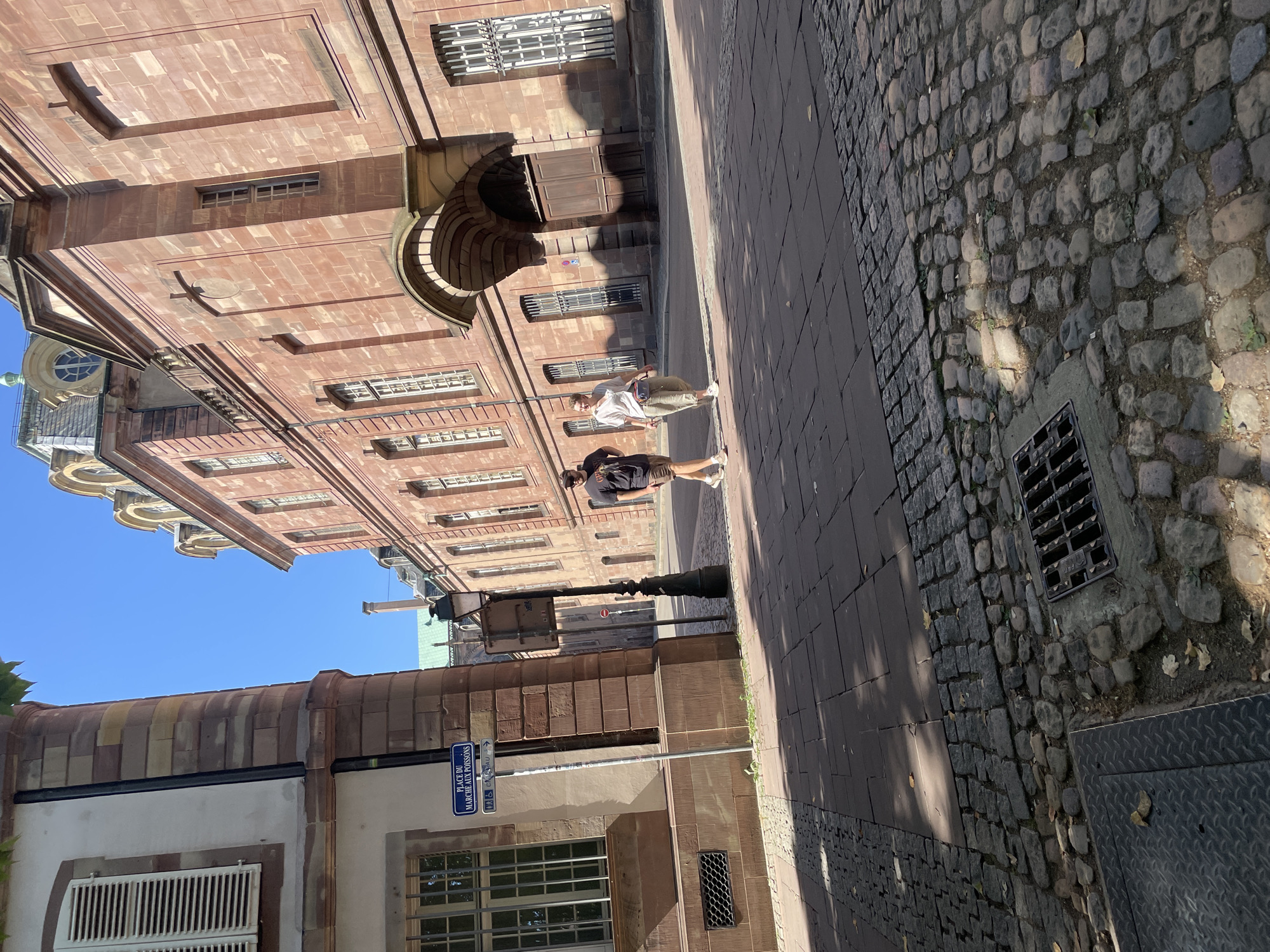 Two individuals are walking down a cobblestone street in Strasbourg, France, on a sunny day. The street is lined with historic, red sandstone buildings. The man on the left is wearing a dark t-shirt and shorts. The woman on the right is wearing a white shirt and light-colored pants, with a small bag slung over her shoulder. A street sign in French, indicating "PLACE DU MARCHÉ AUX POISSONS," is visible on the left side of the street. The ground is paved with a mix of large flat stones and smaller cobblestones. A metal grate, possibly a drain cover, is present on the lower right side of the image. Shadows from the buildings and trees are cast across the street, indicating the time of day is likely late morning or early afternoon.