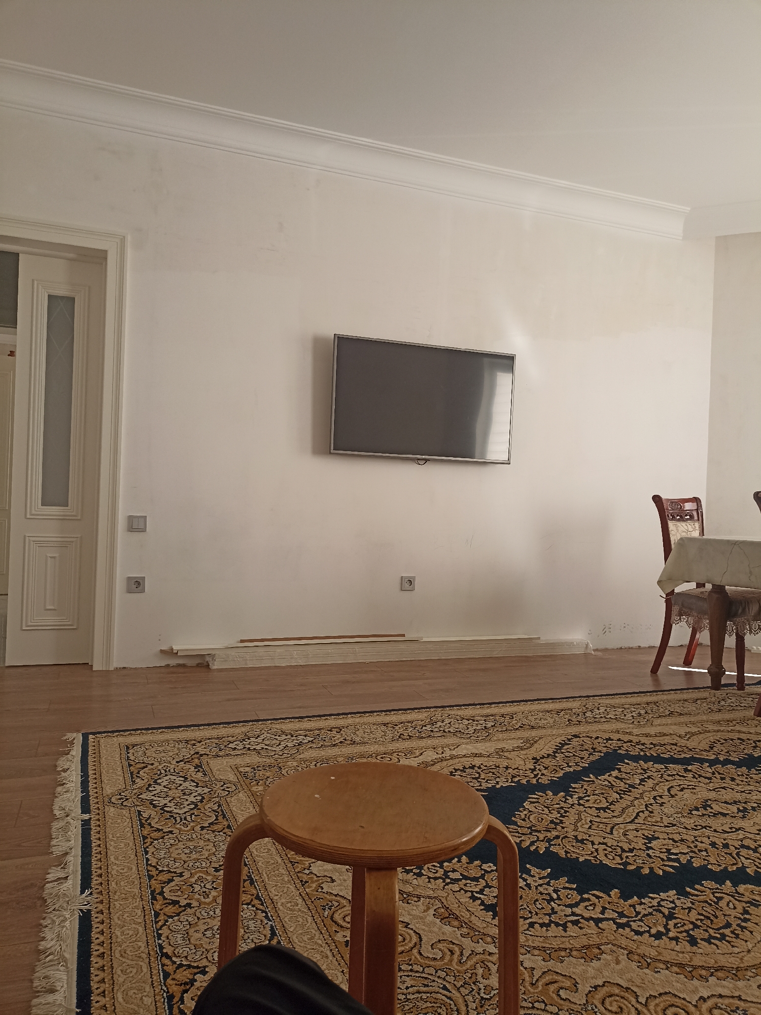 The image depicts an indoor scene, possibly a living room or dining area, located in the city of Izberbash, Russia. The room features a white wall with decorative molding along the ceiling. A flat-screen television is mounted on the wall. To the left, a white door with glass panels and decorative wood trim is visible. Near the door, there are two electrical outlets on the wall. In the foreground, a patterned rug covers a wooden floor. A small, round wooden stool is positioned on the rug. In the background, a wooden chair with a carved back is placed next to a table covered with a patterned tablecloth. Some white trim pieces are stacked on the floor near the base of the wall. The lighting suggests it is daytime. There are no people visible in the image.