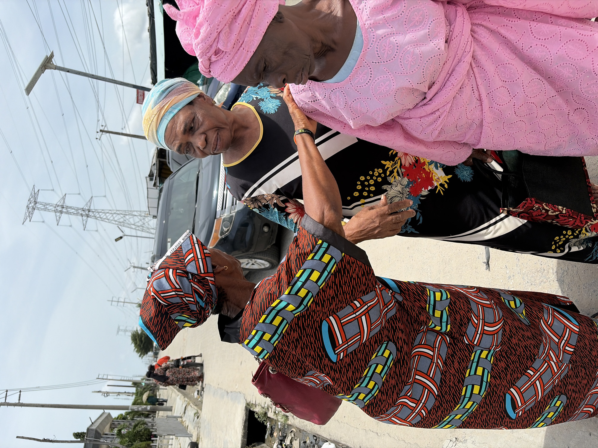 The image captures a scene in Igboefon, Nigeria. Three elderly African women are the primary subjects. One woman is wearing a brown patterned dress and headwrap. Another woman is wearing a black and white top with colorful floral designs and a blue and tan headwrap. The third woman wears a pink lace headwrap and garment. The women are close together, interacting on a sidewalk near a black vehicle and an electrical tower. The background shows urban elements, including a street and buildings. There is some trash and debris on the sidewalk.