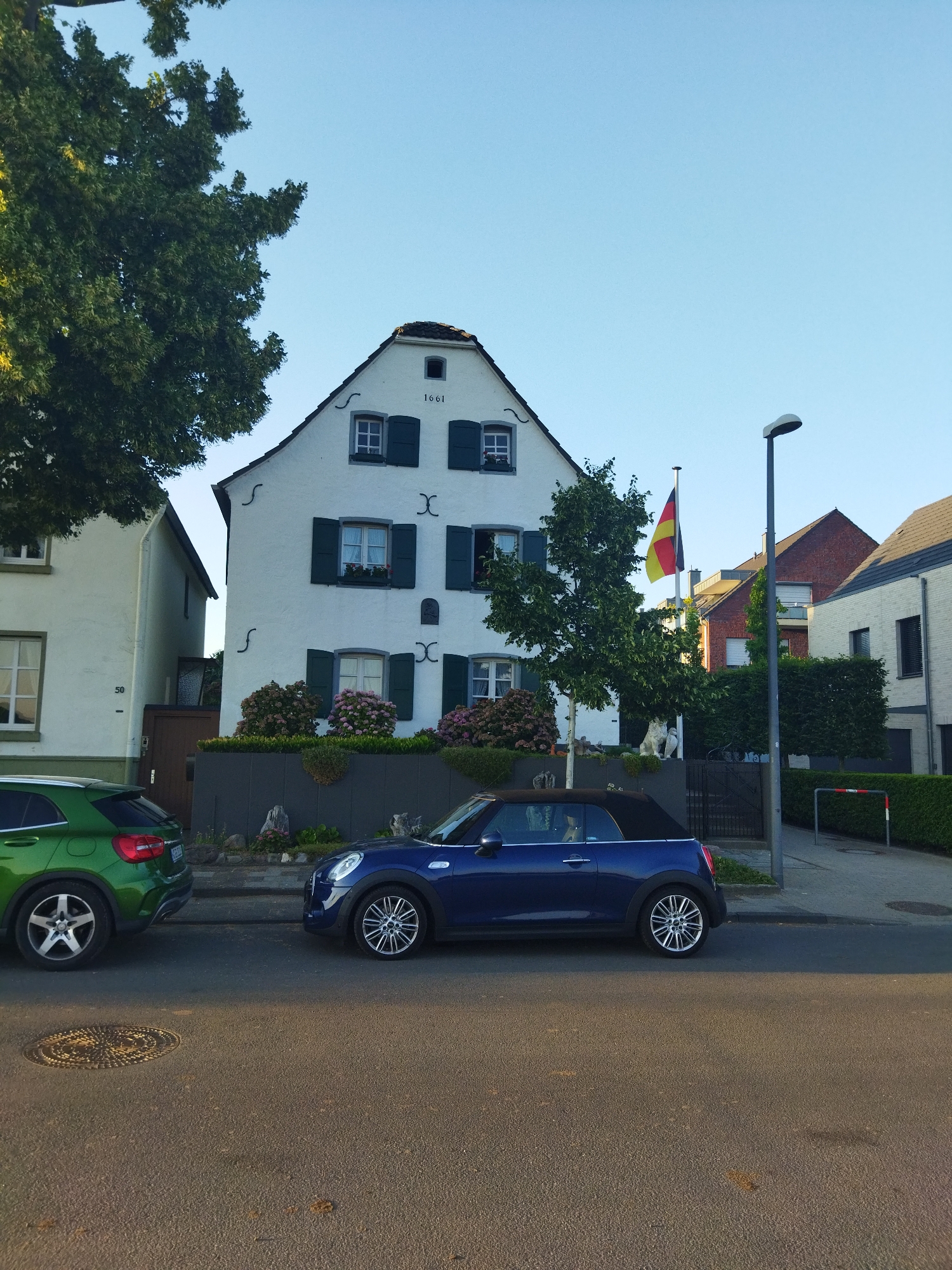 The image shows a street scene in Leverkusen, Germany, with a focus on two parked cars and a white, historic-looking building. A green SUV is parked on the left, and a blue convertible is parked on the right. The white building, which appears to be a house, has dark green shutters on its windows and a date "1661" inscribed above the top-floor windows. A German flag is visible on a flagpole in front of the building. The sky is clear and blue, suggesting it is daytime. The street is paved, and there is a sidewalk with a hedge and a street lamp. Other houses and trees are also visible in the background.