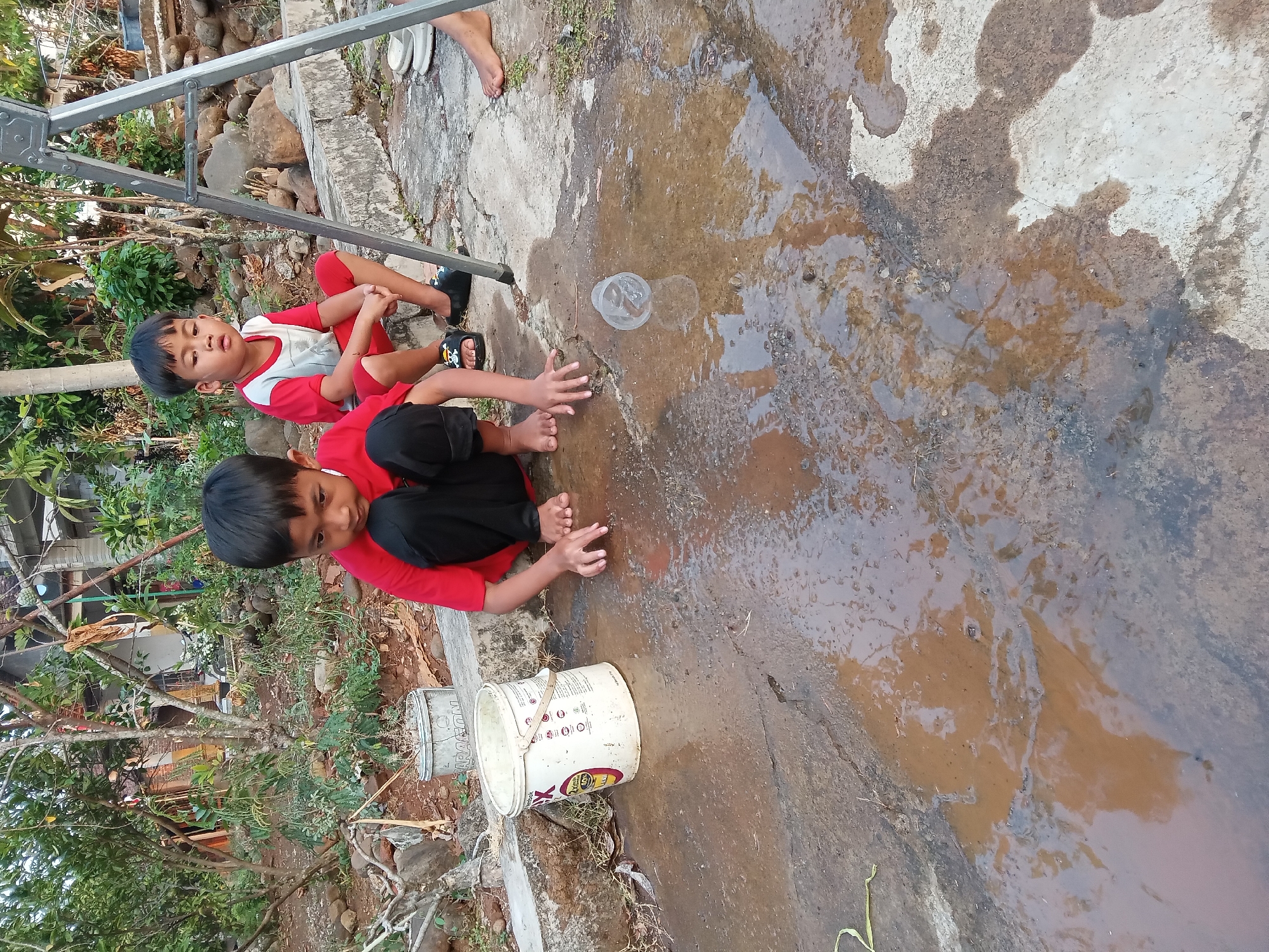 Two young boys are crouching near a puddle of water on a concrete surface. The boy in the foreground is wearing a red shirt and black pants and is reaching his hands into the puddle. The boy in the background is wearing a red and white shirt and red shorts and is sitting with his legs crossed. There is a white bucket with a handle to the left of the puddle. A metal ladder stands to the right of the boys. The background shows trees and a house. The scene appears to be in an outdoor residential area. The location is stated to be Kedunggudel, Indonesia.