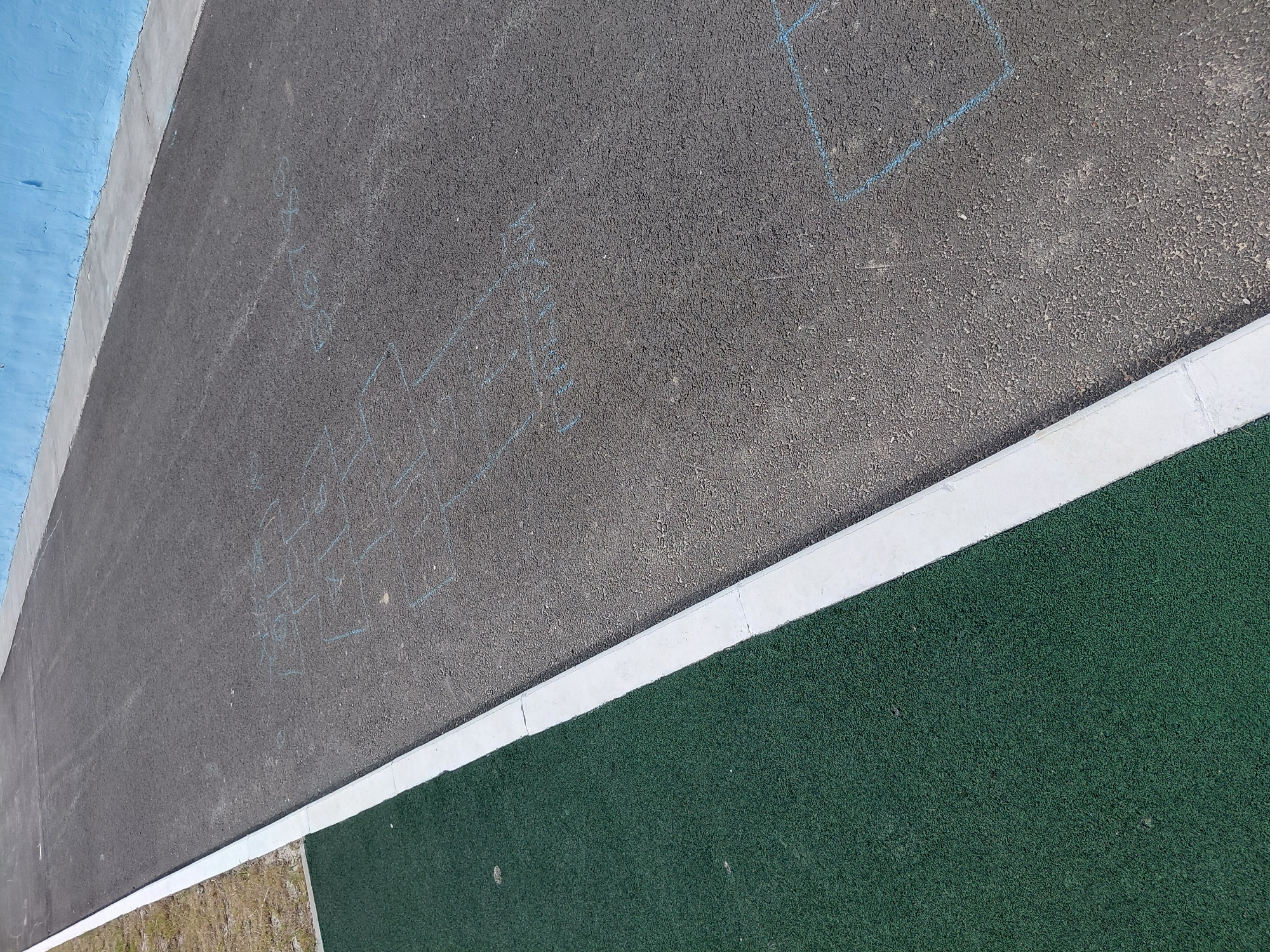 This media file presents an aerial-view image of an outdoor recreational area, likely a playground or sports court, located in Nadym, Russia. The scene is dominated by a vast expanse of dark gray asphalt pavement, showing a textured, granular surface.
To the left, a vibrant blue painted wall rises, bordered by a concrete coping that separates it from the asphalt. A stark white painted curb or dividing line runs diagonally across the lower right section of the image, segmenting the asphalt from a rich dark green, textured surface, which appears to be artificial turf or a rubberized safety surface. In the bottom left corner, a small section of dry, light brown grass and dirt is visible, suggesting a transition to natural ground.
The primary activity suggested in the image is children's play, although no individuals are present. This is evidenced by multiple drawings sketched with light blue chalk on the asphalt. The most prominent drawing is a complex, non-standard hopscotch-like grid featuring various interconnected boxes and lines. Within some of these boxes, faint and irregularly drawn letters or symbols can be discerned, such as what appears to be "D", "K", and "L", though their complete interpretation is challenging due to their faded nature. Another simpler, square-shaped chalk outline is visible further up and to the right on the asphalt.
Notable details include the bright, clear lighting, indicating it is daytime, likely sunny, given the sharp contrast and lack of long shadows. The overall impression is one of a quiet, prepared space awaiting or having recently hosted recreational activities.