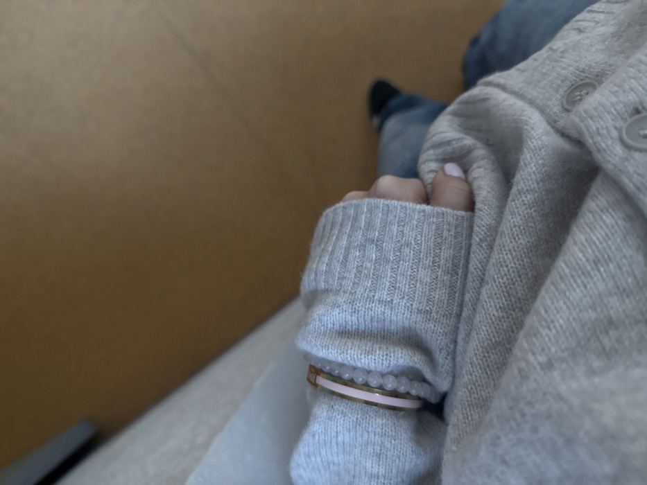 A top-down shot features a person's clothed arm, adorned with bracelets, resting against a light grey cardigan and blue jeans. A glimpse of a black shoe is visible. The cardigan has visible buttons. The bracelets are thin, one gold with a pink band and another string of light-colored beads. The hand partially obscured by the sleeve, with light pink nail polish visible. The setting appears to be indoors, with a tan wall in the background. A white surface is partially visible below the arm.