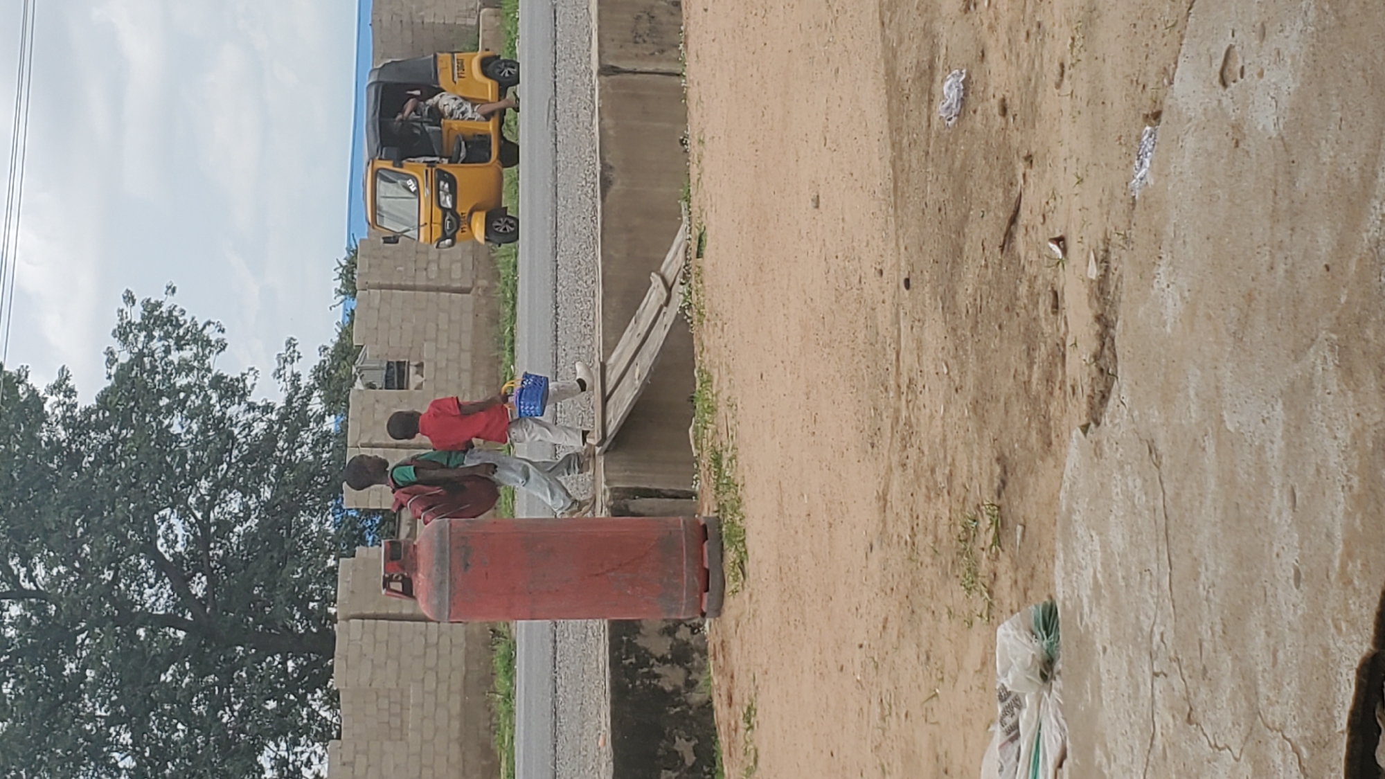 In Jalingo, Nigeria, two young boys are walking on a dirt path next to a gray asphalt road. The boy on the left wears a green shirt and a maroon backpack, while the boy on the right is in a red shirt and white pants, carrying a blue basket. A large red propane tank stands to the left of the boys. Across the road, a yellow and black auto-rickshaw is parked, with a person visible in the driver's seat and another person entering the vehicle. The background consists of a low, light-colored brick wall under a cloudy sky, with a large tree to the left. A wooden ramp spans a concrete ditch in front of the road. The foreground is composed of sandy ground with some patchy green vegetation and scattered debris, including a white plastic bag.