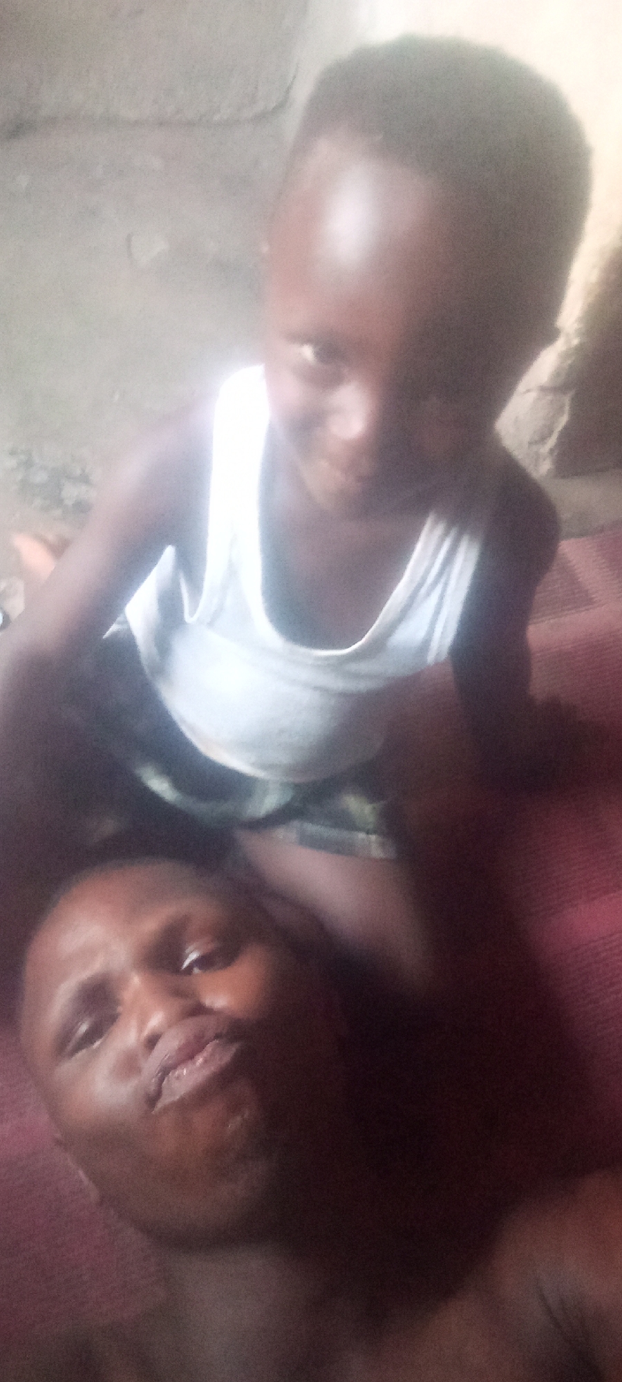 A man lies on his back on a patterned maroon and grey mat, possibly a blanket, with a young child perched on top of him. The child is wearing a white tank top. Both the man and the child appear to be looking at the camera. The background is out of focus, but suggests a simple interior with a rough wall. The image provides no evidence to confirm it was taken in Sabon Tasha, Nigeria.