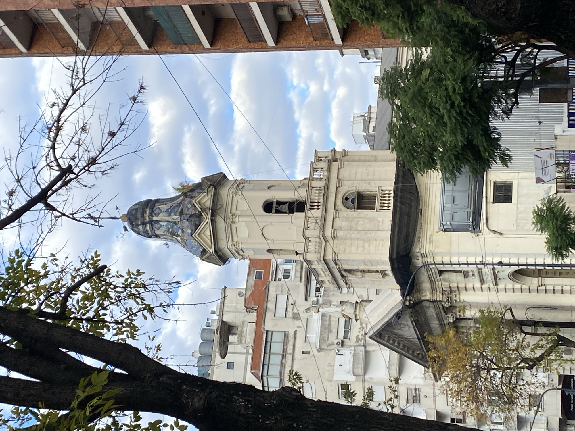 The image is a low-angle shot of a church in Buenos Aires, Argentina. The church features a distinctive dome with blue and white tiles. Its façade is off-white with classical architectural details. The shot is framed by trees with bare branches in the foreground, suggesting a possible fall or winter season. A building with rectangular architectural details is visible at the top of the frame. The sky is overcast with clouds. Other buildings of different heights and styles surround the church.