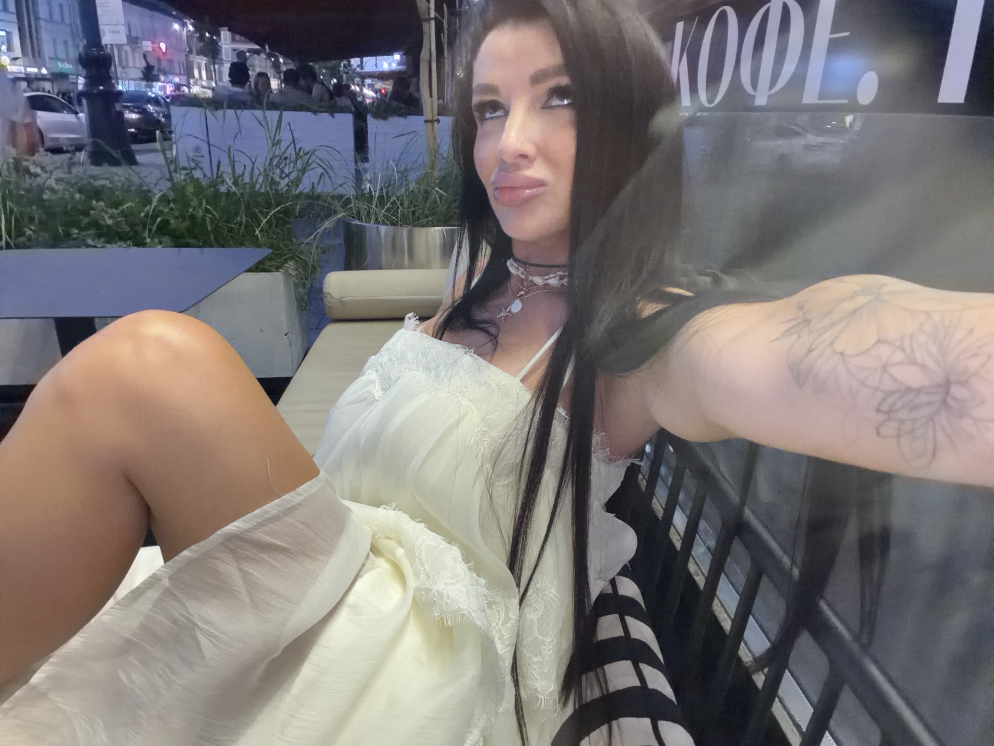 A woman with long dark hair and prominent makeup is seated at an outdoor cafe. She is wearing a white lace-trimmed dress and a choker necklace with a shell pendant. Her left leg is crossed over her right, and her right arm is extended, with a tattoo visible on her forearm. The background shows a street scene with cars, buildings, and greenery. A sign with Cyrillic lettering, possibly indicating "Coffee," is visible. The setting appears to be nighttime or dusk. The location is identified as Novaya Usman', Russia.