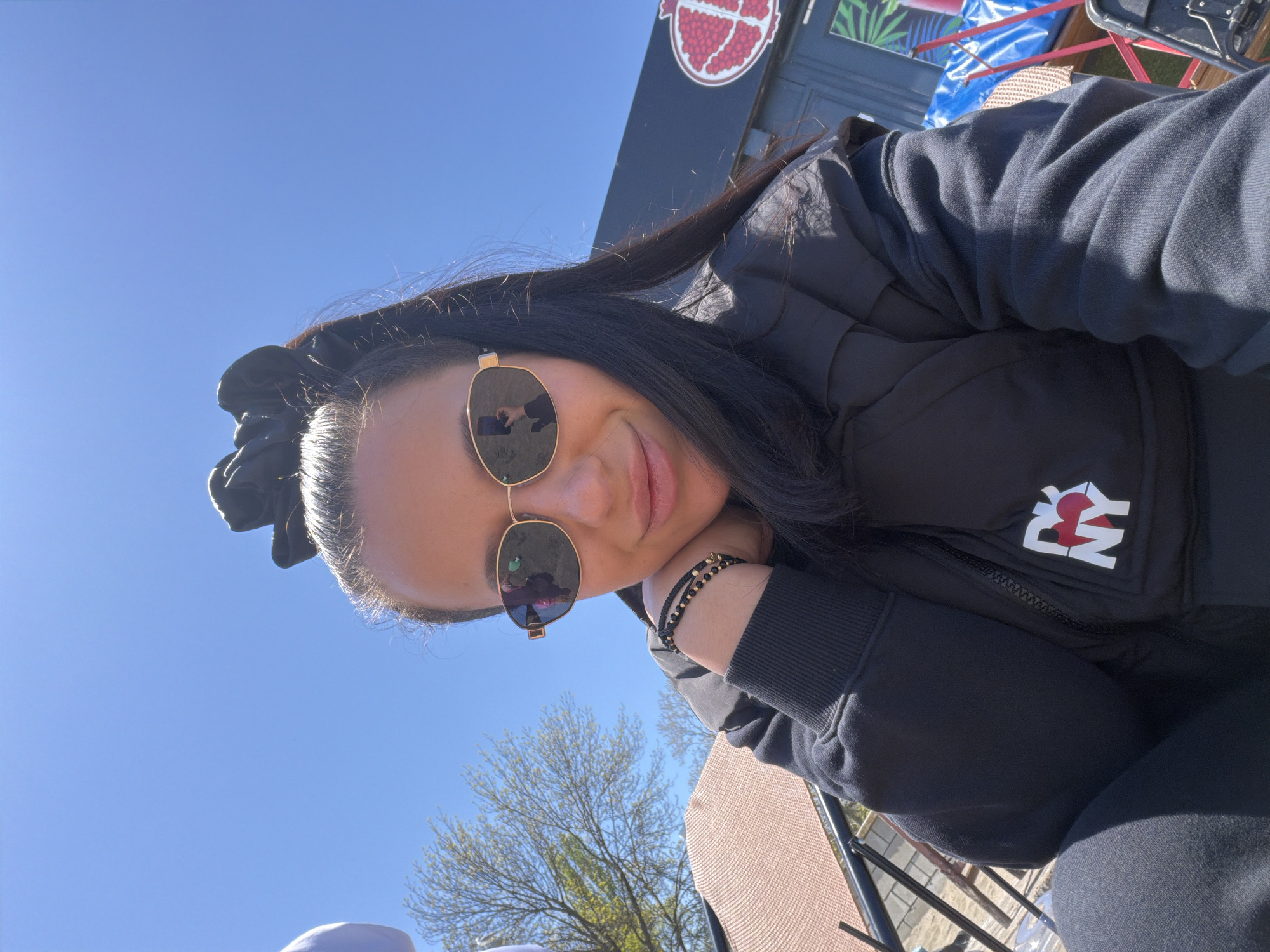A person wearing sunglasses and a black jacket with a "NY" logo is sitting outdoors. The person has dark hair pulled back with a black scrunchie. The background features a blue sky, trees, and part of a building with a pomegranate logo. The location is Unknown, Unknown.