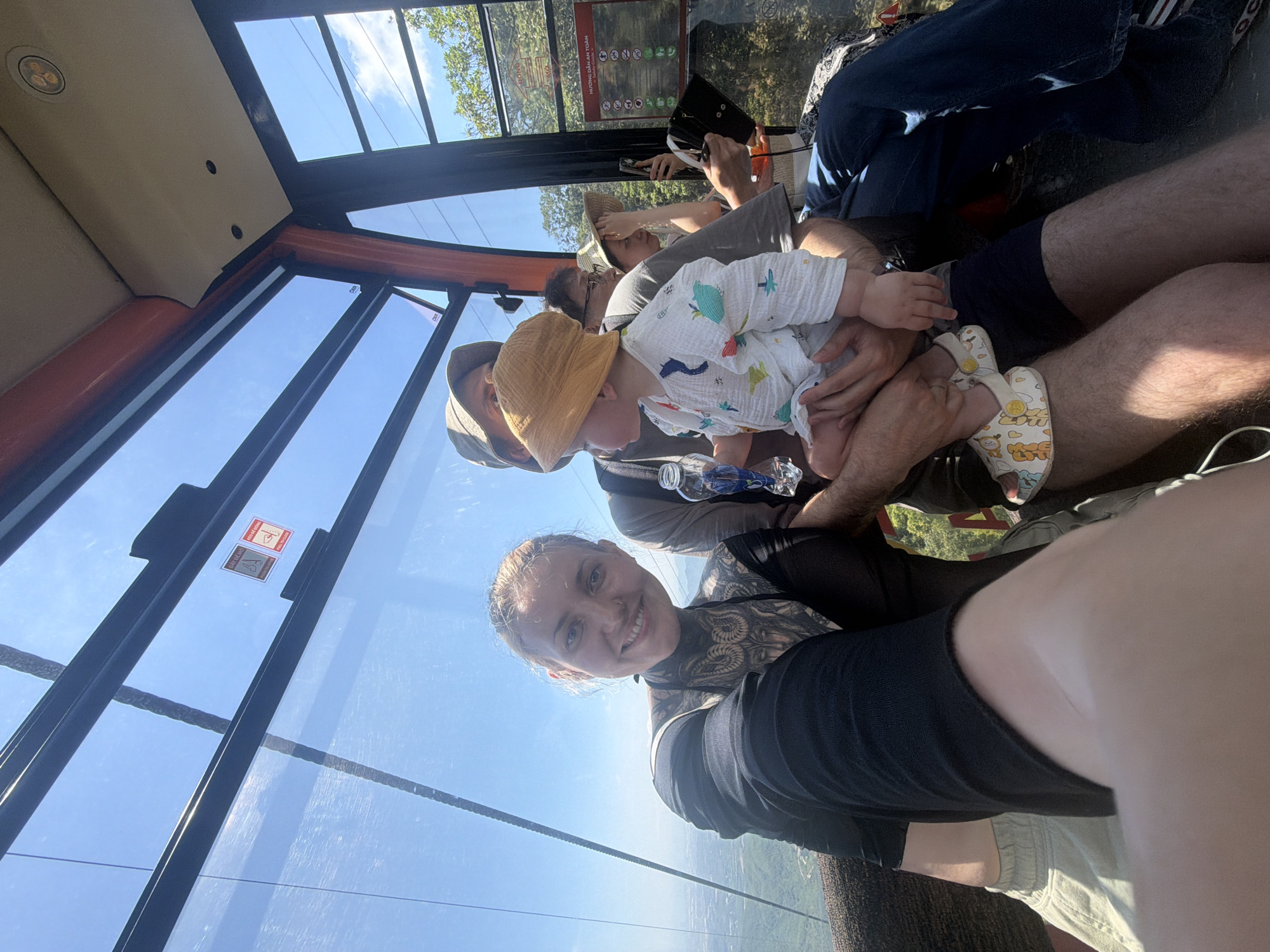 A woman is taking a selfie in a cable car with glass windows, likely in Đà Nẵng, Vietnam. She is wearing a black top and has tattoos on her neck and chest. In the cable car, a man is holding a baby wearing a yellow hat and a patterned onesie. Other people are visible in the background, including a man taking photos with a camera. The cable car offers a view of the landscape below.