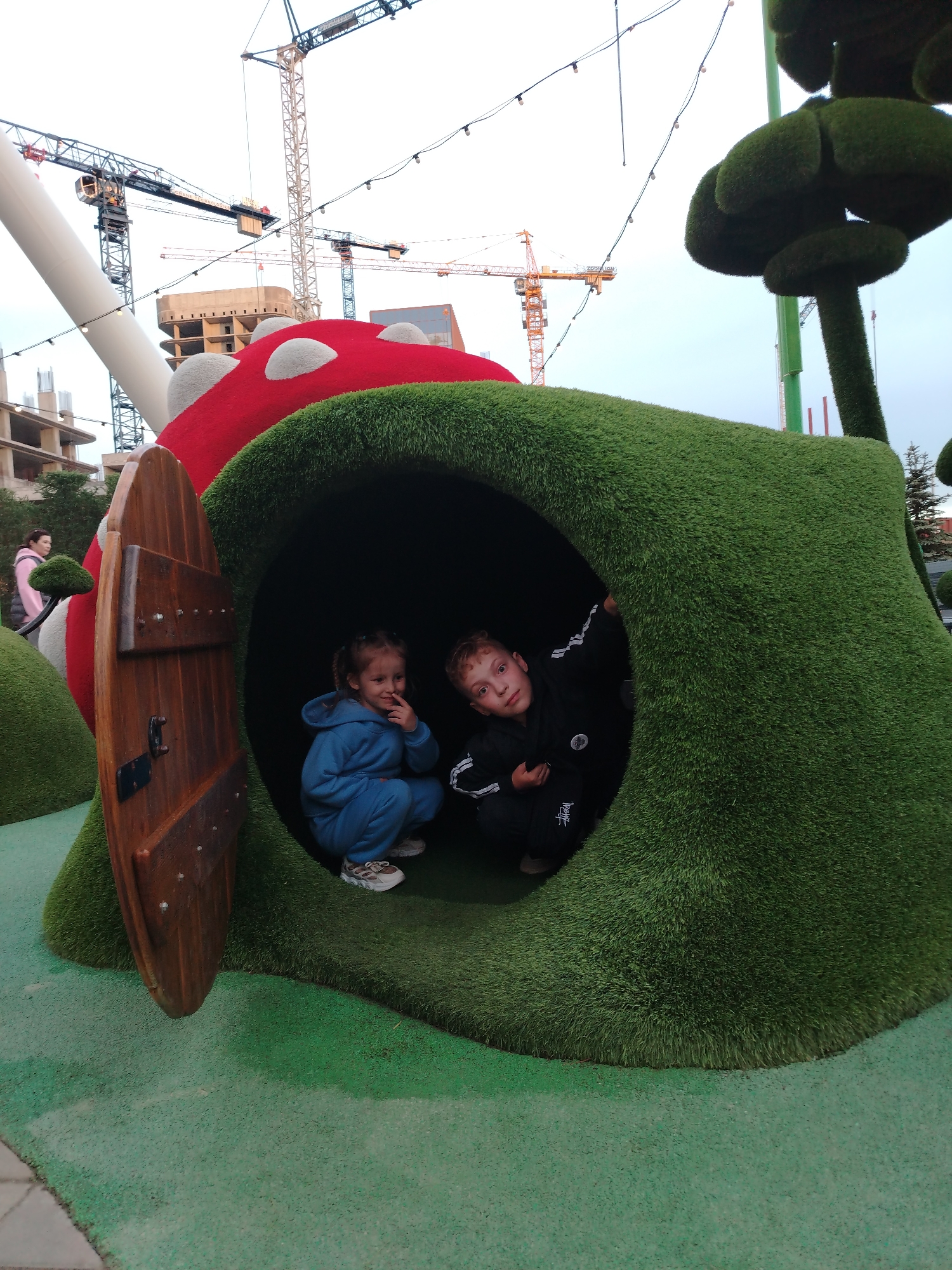 Two children are crouching inside a play structure resembling a green, grassy hill with a red mushroom on top. One child wears a blue tracksuit, and the other wears a black tracksuit. A wooden door is slightly ajar on the left side of the structure. In the background are construction cranes and buildings, suggesting an urban environment. A woman in a pink jacket is partially visible in the background to the left. The location is in the city of Kazan, Russia.