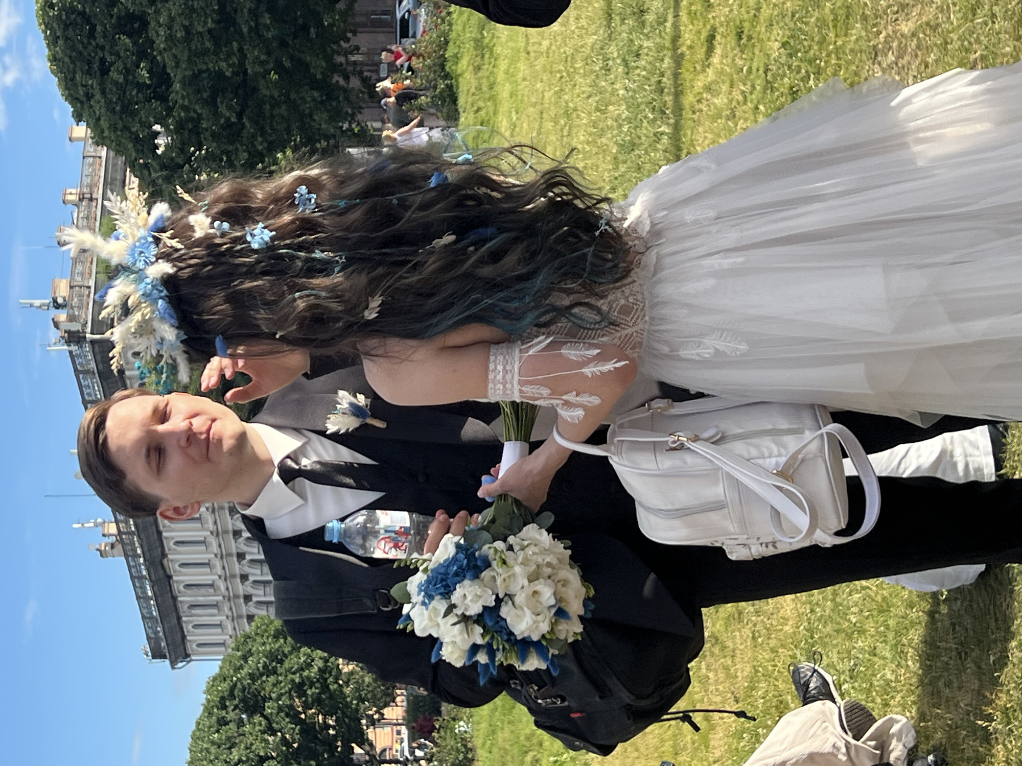 The media file shows a wedding couple outdoors, with the groom in the foreground and the bride partially visible behind him. The groom is wearing a black suit, white shirt, and black bow tie, with a boutonniere on his lapel. He is looking towards the camera with a slight smile. The bride is wearing a white wedding dress with intricate lace detailing on the bodice and sleeves. Her long, dark hair is adorned with a floral and feather crown in shades of white and blue. She is holding a bouquet of white and blue flowers.
A white handbag with gold hardware is hanging from her arm. There is a bottle of water and a backpack visible near the couple. The background consists of a lush green lawn, large trees, and a building with classical architecture, suggestive of a park or a historical estate. The sky is blue with some clouds, indicating a pleasant, sunny day. The scene appears to be a wedding ceremony or reception taking place in Saint Petersburg, Russia, given the architectural style of the building. The overall mood is festive and joyful.