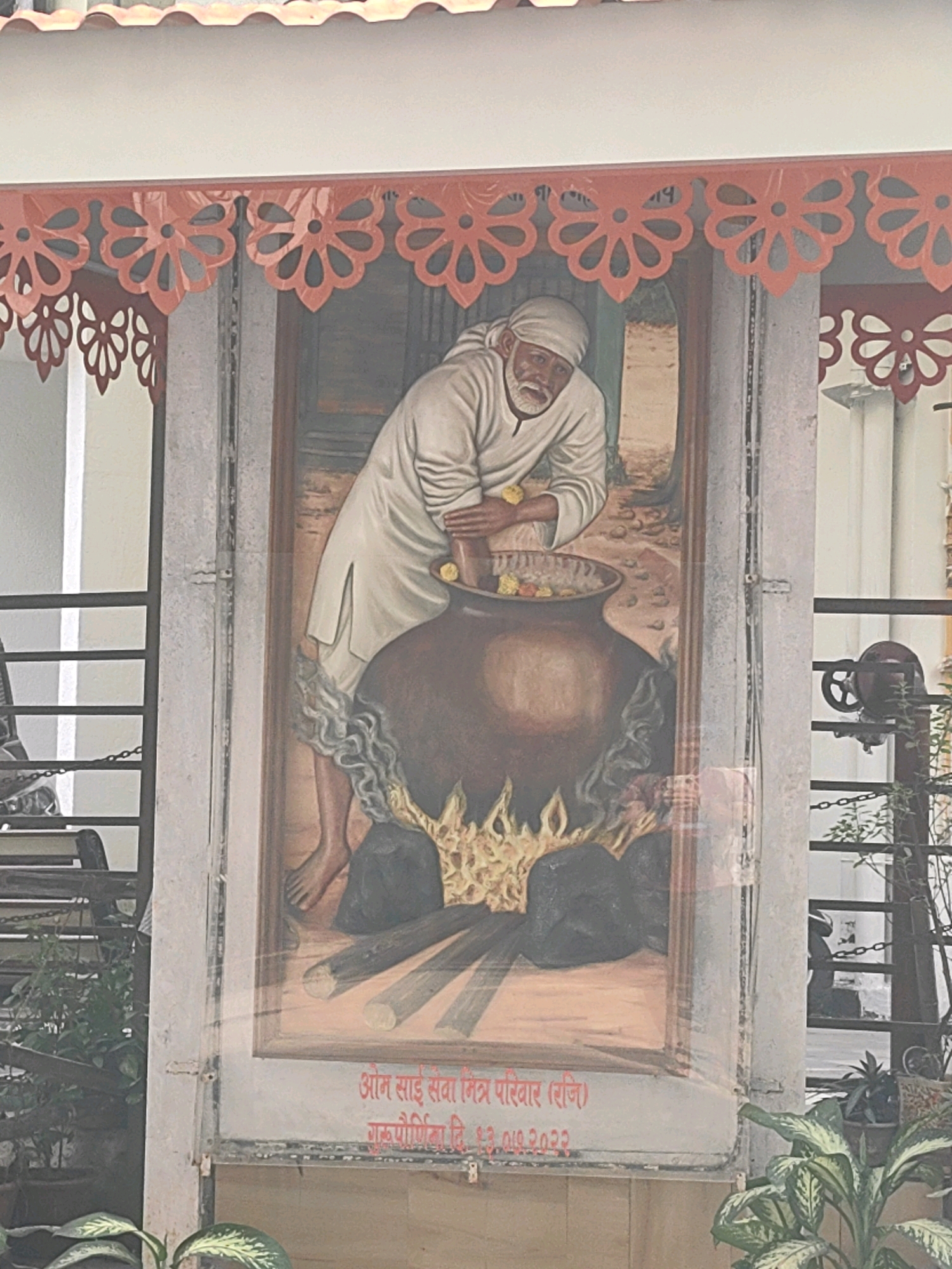 The image shows a mural of a man dressed in white robes, with a white turban, stirring a large pot over a fire. The mural is decorated with red floral cutouts along the top edge. Below the mural, there is text in red. The mural is mounted on a grey, textured wall with vertical metal fixtures. In the background, there are metal railings, some greenery, and a glimpse of a building. The scene appears to be outdoors. The text below the mural, "ॐ साई सेवा मित्र परिवार (रजि.)" and "गुरुपौर्णिमा दि. ०३.०७.२०२३" suggests this is likely located at a religious or community center in Mumbai, India, celebrating Guru Purnima.