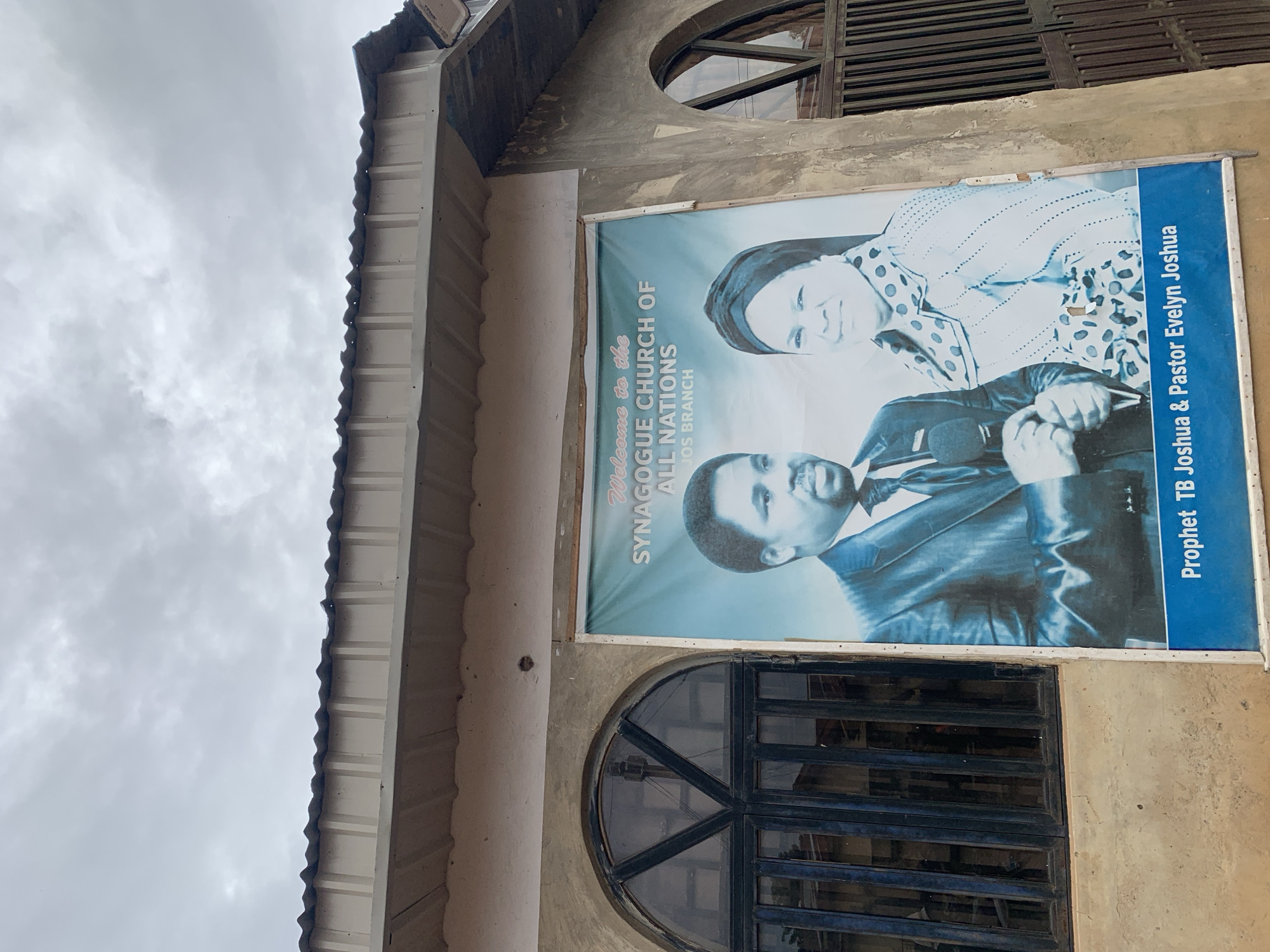A large banner for the Synagogue Church of All Nations, Jos Branch, is displayed on the exterior of a building in Rafin Par, Nigeria. The banner features portraits of Prophet TB Joshua and Pastor Evelyn Joshua, with text indicating the church name and their titles. The building has a corrugated metal roof and two arched windows with dark-colored frames. The wall is made of concrete and appears weathered. The sky is overcast with clouds.