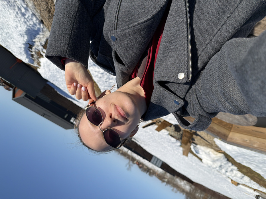 A female individual is depicted in a close-up, upward-angled outdoor photograph, taken on a clear, sunny day in Shipulino, Russia. She has light skin, dark hair pulled back, and wears gold-framed sunglasses with dark lenses. Her expression is neutral with a slight upward turn of the lips. She is dressed in a dark grey, textured, long-sleeved winter coat featuring a zippered front and silver-toned snap buttons on the lapels and shoulders, over a visible red garment. Her right hand is raised to the side of her head, with fingers positioned near her temple; her fingernails are painted light pink with small white polka dots. A silver-toned chain-link earring is visible on her right ear. The background shows ground partially covered in snow, with exposed patches of earth and various wooden structures, possibly benches or decking, to the mid-left. Further in the background, bare trees or bushes line the horizon to the left, while a dark, rectangular building or structure is visible to the upper right. The sky is clear and bright blue.