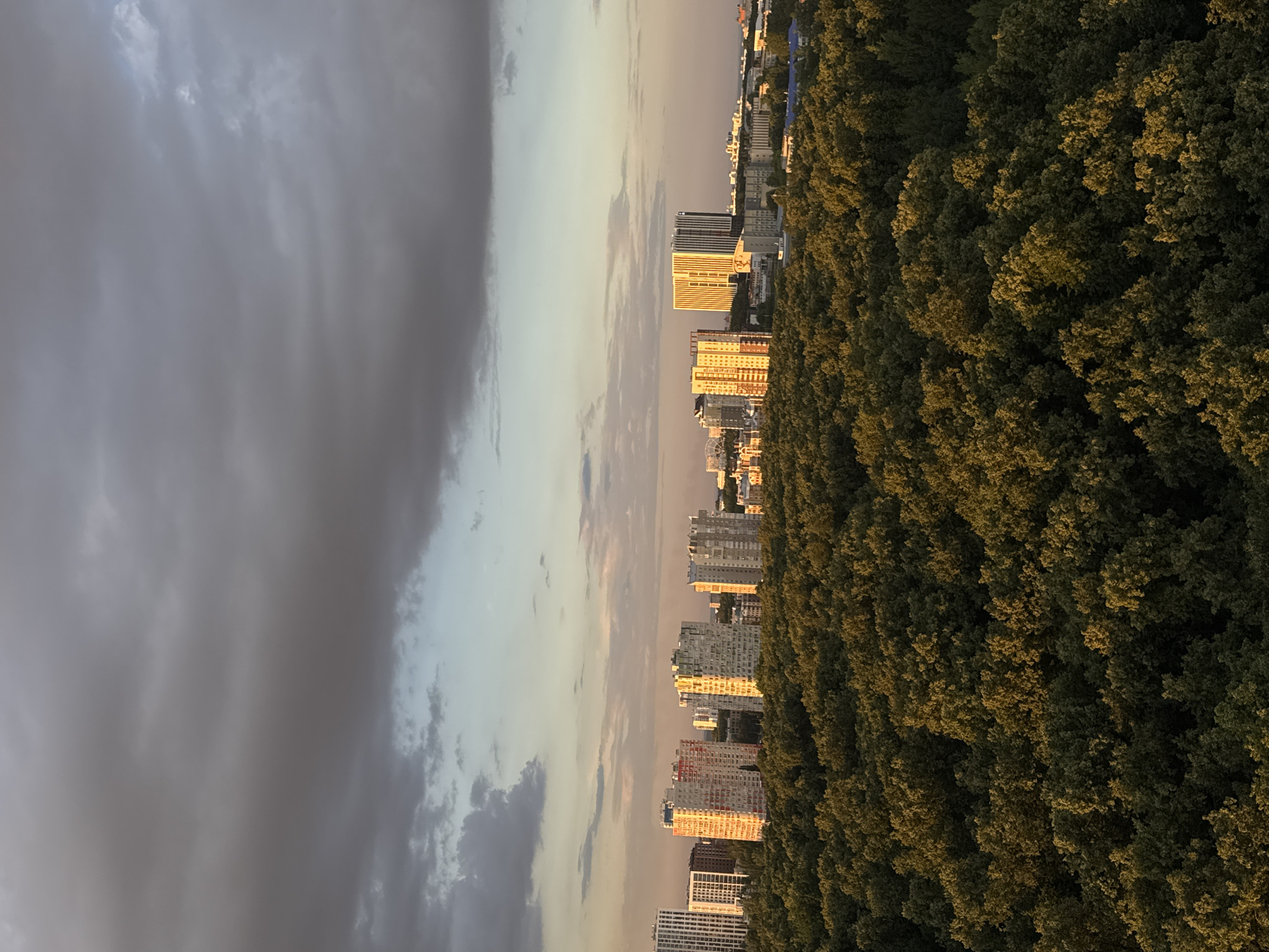 The image captures a panoramic view of Ufa, Russia, at sunset. In the foreground, a dense forest stretches across the frame, its lush green canopy illuminated by the warm, golden light of the setting sun. Beyond the trees, a cluster of modern high-rise buildings punctuates the skyline. These buildings exhibit varied architectural styles, with some displaying colorful facades that catch the sunset's glow. The sky above is a dramatic canvas of dark, heavy clouds interspersed with patches of pale blue and hints of orange and pink, suggesting the transition from day to night. The overall scene conveys a sense of urban development encroaching upon a natural landscape, set against the backdrop of a striking sunset.