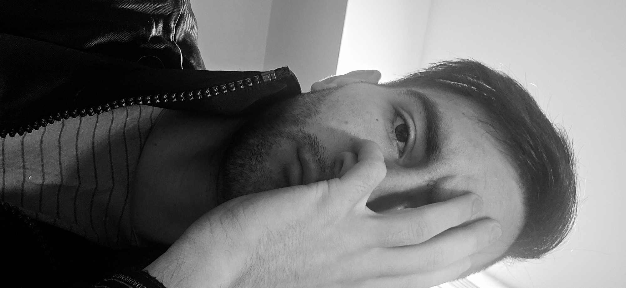A black and white close-up photograph depicts a man. His right eye is visible. His right hand is covering the lower part of his face. He has short dark hair and a beard. He is wearing a striped shirt and a jacket with a zipper. The setting appears to be indoors. The photograph does not provide information to determine the location in Plovdiv, Bulgaria.