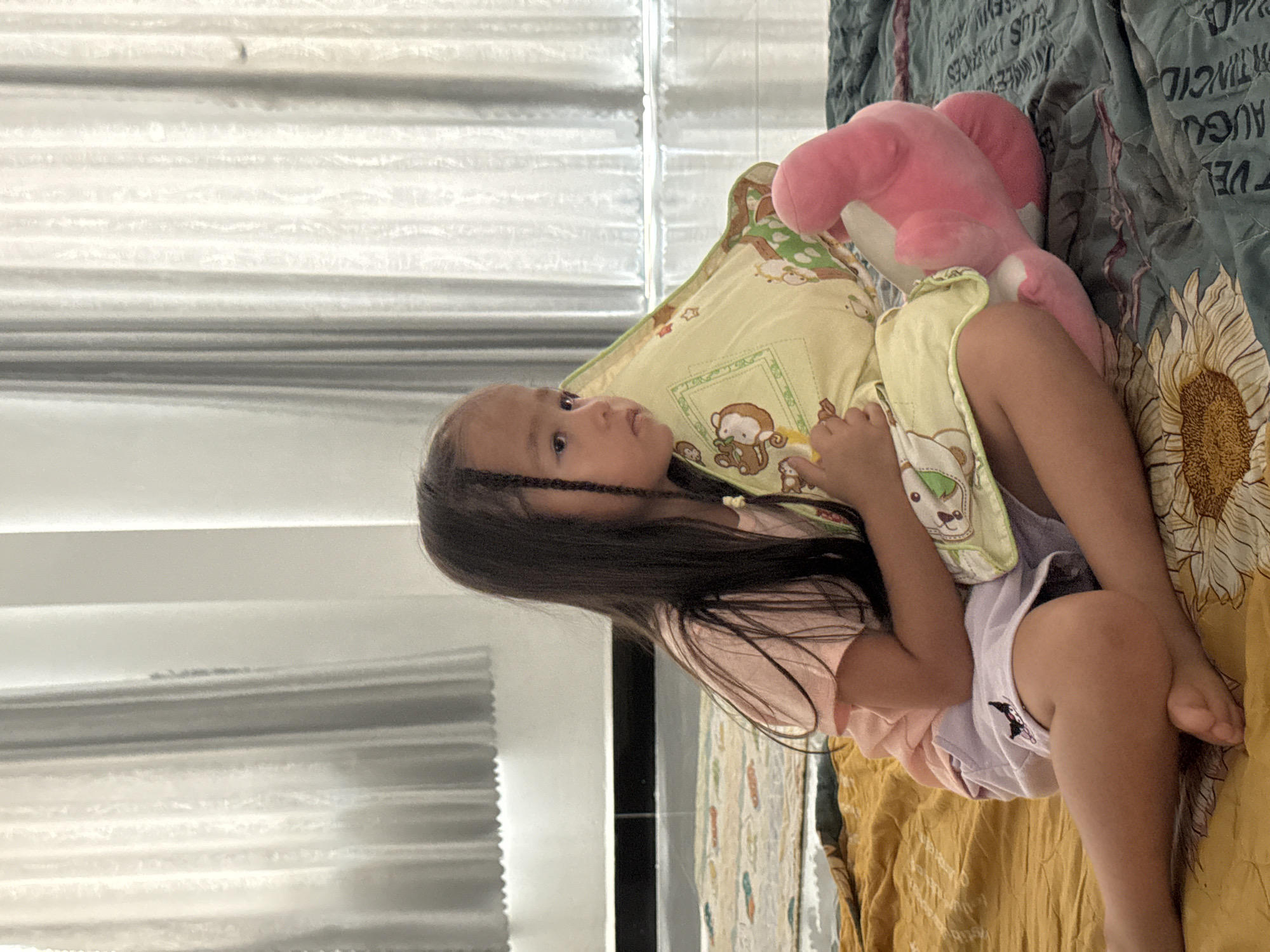 A young girl with long, dark hair, braided on one side, lies on a bed with a patterned quilt and a yellow pillow decorated with cartoon monkeys. She is wearing a light pink shirt and light purple shorts. A pink stuffed animal rests near her head. The location appears to be indoors, possibly a home, with a window featuring horizontal blinds visible in the background. No further details connect the image specifically to the city of Mũi Né, Vietnam.