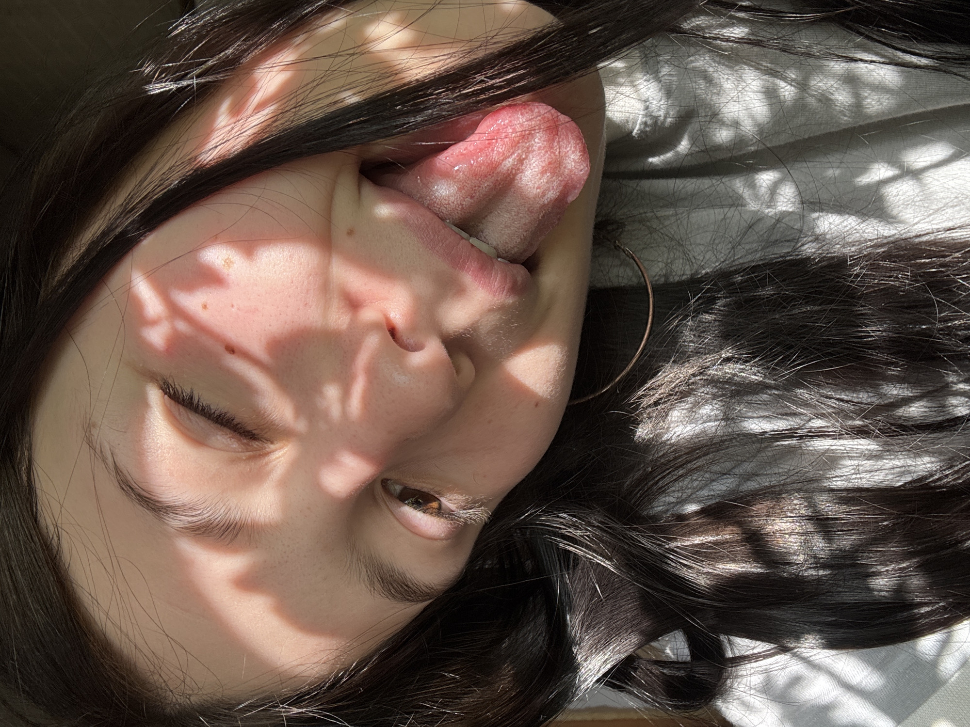 The image shows a person's face close-up, tilted to the left. The person has dark hair and light skin, with their tongue sticking out. Light and shadow patterns fall across their face. They appear to be indoors, with light coming through what may be foliage. The environment lacks features indicating the location of Фоки, Russia. The person's eye is partially closed.