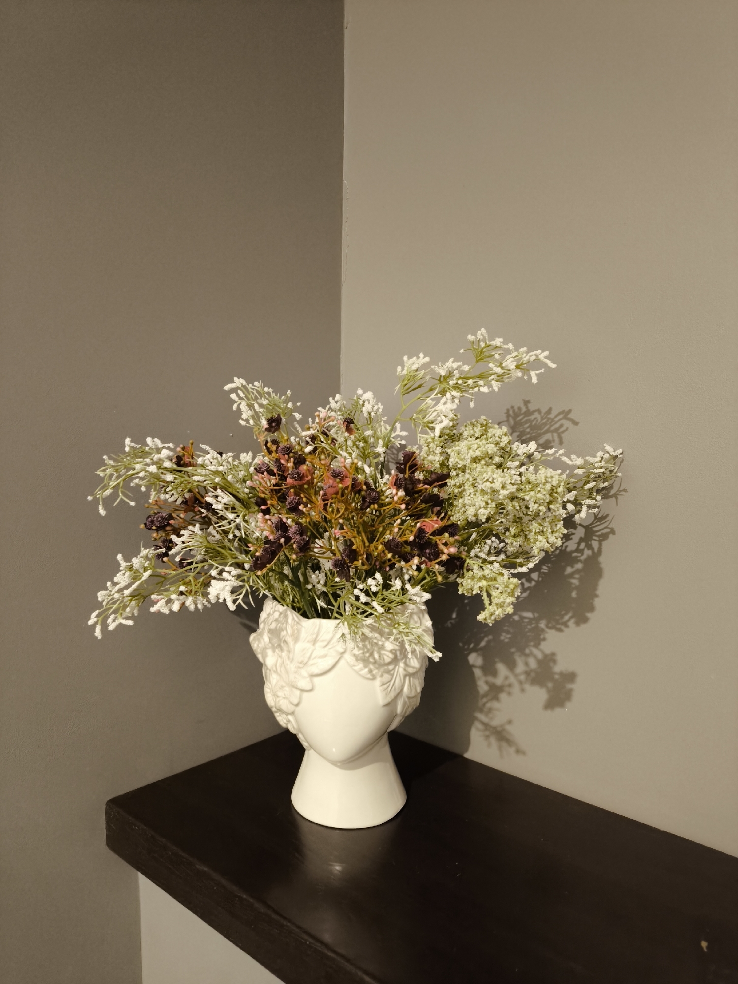 A still-life composition shows a white, head-shaped vase filled with artificial flowers. The flowers are a mix of white, green, brown, and dark purple hues. The vase is positioned on a dark wooden surface, likely a shelf or table. The background consists of two walls meeting at a corner, both painted in a muted gray or beige tone. The lighting appears to be artificial, casting shadows of the flowers onto the right wall. There is no indication of this being located in Smolensk, Russia or any other specific location.