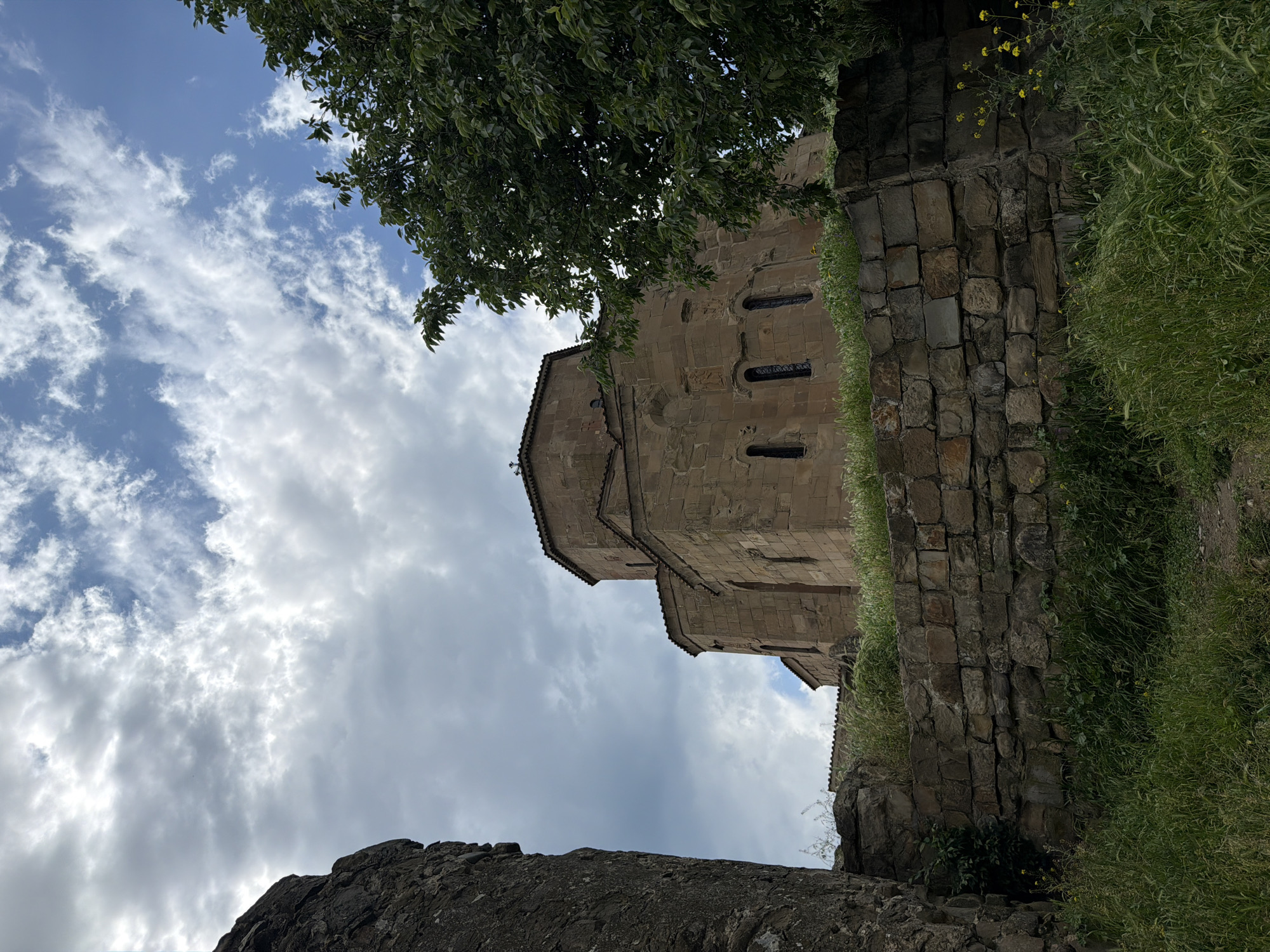 The image depicts Jvari Monastery, located near Mukhatgverdi, Georgia, from a low-angle perspective. The stone building of the monastery is visible against a backdrop of a cloudy sky. Part of a tree's foliage is positioned above the monastery structure, partially obscuring it. A rough stone wall covered with grass and some yellow flowers flanks the right side of the monastery. In the foreground, at the bottom of the frame, a portion of another stone structure is visible.