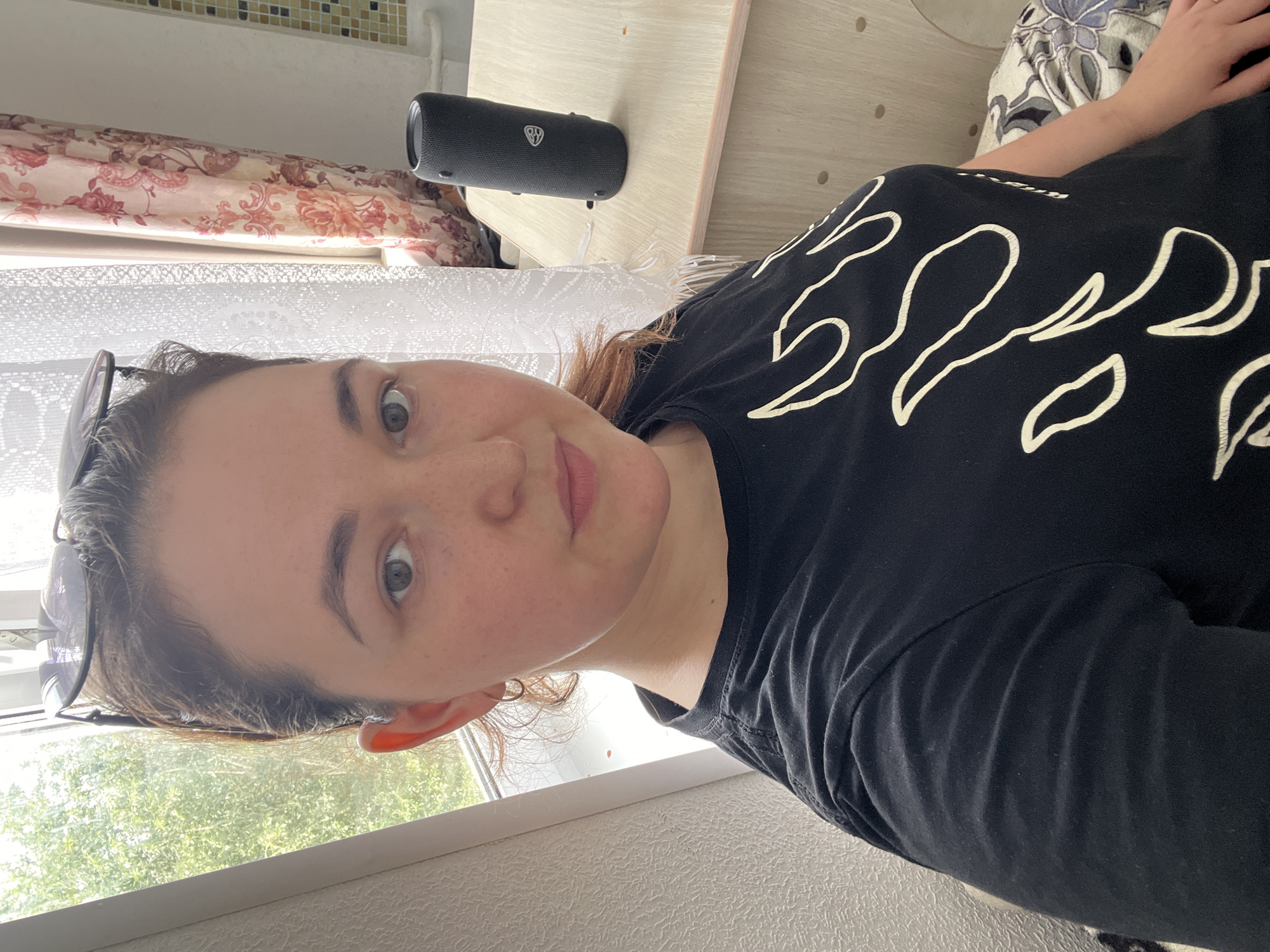 A young woman with brown hair and light skin is taking a selfie indoors. She is wearing a black t-shirt with a white flame-like design. Sunglasses are perched on top of her head. Her face is looking directly at the camera, and she has a neutral expression. To her right, a black cylindrical speaker sits on a wooden desk next to a window with lace curtains and a floral pattern. The background suggests a domestic setting, possibly an apartment in Chelyabinsk, Russia. Sunlight streams in from the window, illuminating the scene.