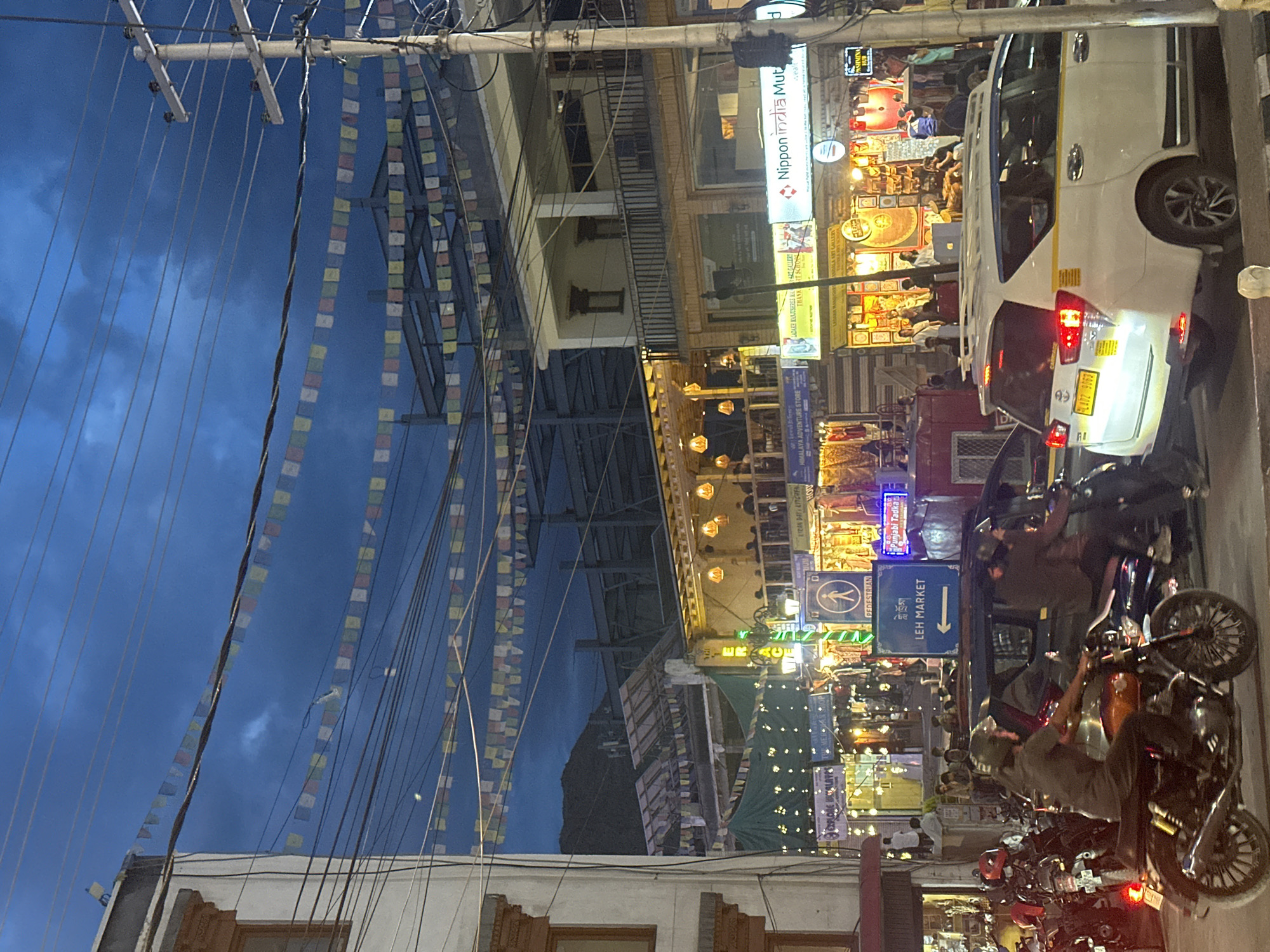 The image depicts a street scene in Leh, India, likely during the evening. The sky is a deep blue, indicating dusk or a cloudy evening, with prayer flags strung across overhead wires. Several multi-story buildings line the street, featuring shops and restaurants with illuminated signs and decorative lighting. One sign clearly reads "LEH MARKET" with an arrow pointing left.
In the foreground, vehicles are present. A white SUV taxi with the license plate "PL47Z 9062" is parked on the right side of the road. To its left, a dark-colored car is visible. In the left foreground, a person is riding a motorcycle, wearing a helmet and a dark-colored shirt. Another motorcycle is parked nearby. Several other individuals are visible in the background, some appearing to be customers or staff in front of the shops. The street is lined with various businesses, including what appears to be an adventure store and art galleries. The overall atmosphere suggests a bustling market area with a blend of local and commercial activity.