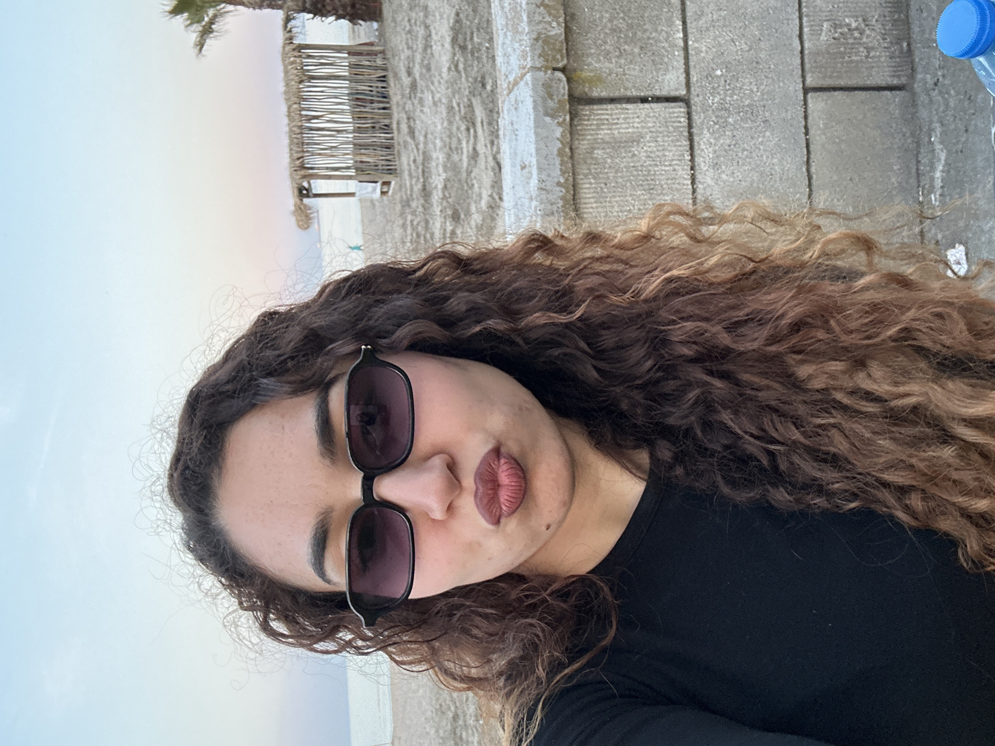 This image, captured in what appears to be a vertical orientation but displayed rotated counter-clockwise, features a young woman taking a selfie in an outdoor setting, likely in Burgas, Bulgaria. The central subject is a woman with long, dark, and voluminous curly hair. She wears dark-framed rectangular sunglasses with dark lenses. Her expression is characterized by pursed lips, highlighted with a reddish-pink matte lipstick, giving a stylish and playful vibe. She is dressed in a simple black long-sleeved top.
The background suggests a coastal or beach environment. Behind the woman, a sandy area is visible, bordered by a rustic wooden slatted structure, possibly a fence or part of a beachside establishment. A portion of a palm tree is visible towards the top. The foreground and area immediately adjacent to the woman feature grey concrete or stone pavement. A plastic water bottle with a blue cap is partially visible in the upper right section of the image (when viewed upright). The soft, muted tones of the sky indicate either an early morning sunrise or a late afternoon sunset, or perhaps an overcast day, contributing to a tranquil atmosphere. There is no discernible text within the image.