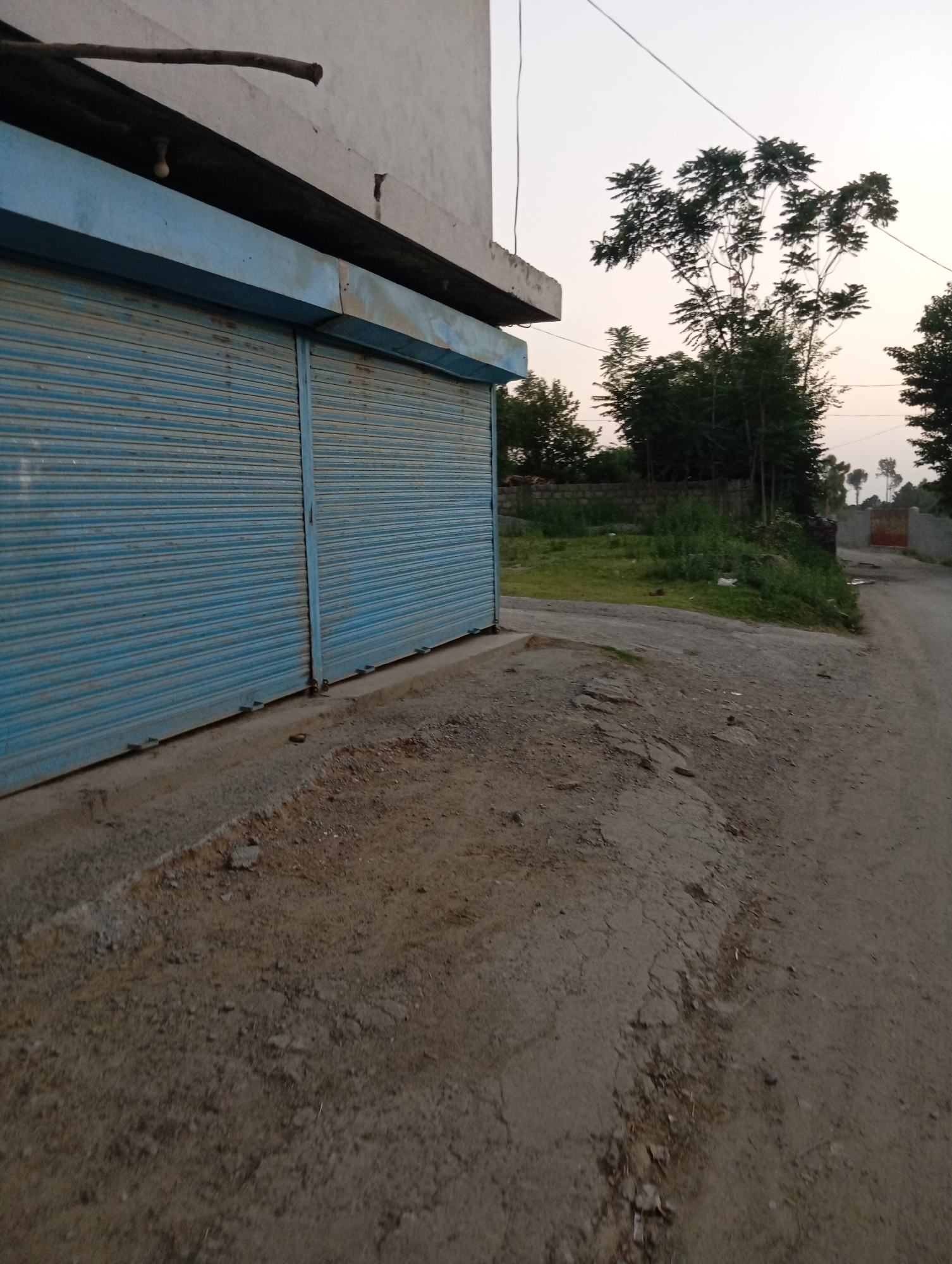 The image depicts an outdoor scene in Lassan, Pakistan. In the foreground, a closed blue corrugated metal shutter covers the entrance to what appears to be a shop or building. The shutter is weathered and shows signs of rust. Adjacent to the shutter, a dirt path or road curves to the right. The ground is uneven and covered with gravel and dirt.
In the background, there is a patch of green grass and vegetation, with some trees visible. A low stone wall borders some of the grassy area. Further in the distance, more trees can be seen, and the sky is a pale, overcast hue, suggesting it might be dawn or dusk. There are no people or animals visible in the frame. The overall impression is one of a quiet, perhaps slightly neglected, rural or semi-urban setting.