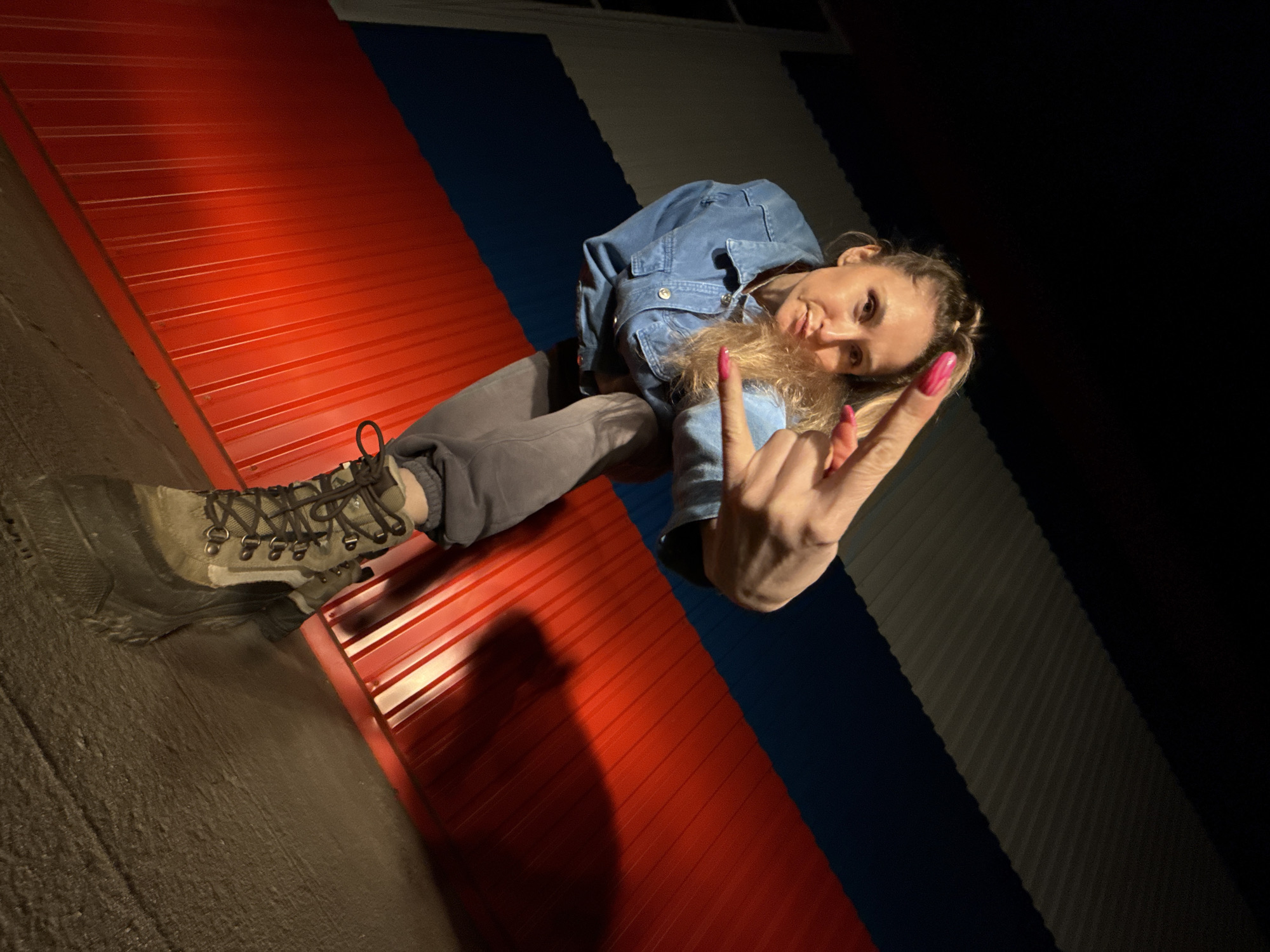 A person wearing a denim shirt and gray pants sits on a tricolor corrugated metal surface, appearing to represent the flag of Russia. The colors are red, white, and blue. The person wears a boot with tied laces on one foot and makes a "rock on" hand gesture. The background appears to be dark. The image appears to be taken at night in an outdoor setting. There is no verifiable indication in the image of the location being Chaikovsky, Russia.
