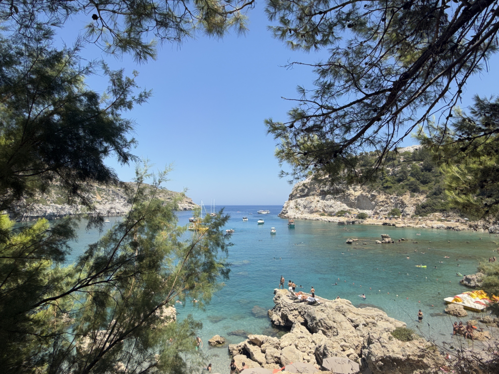 A scenic bay in Rhodes, Greece, is captured on a bright, sunny day. The clear, turquoise water teems with people swimming, sunbathing on rocks, and enjoying various water activities. Several boats, including sailboats and smaller vessels, are anchored in the bay. The shoreline is dotted with rocky outcrops and lush green vegetation, with steep, rocky cliffs framing the scene.
In the foreground, pine branches and foliage partially obscure the view, adding a natural, framed perspective to the idyllic setting. On a prominent rocky ledge in the middle ground, several individuals are relaxing, some sunbathing on lounge chairs and others standing and observing the bay. More swimmers and people wading in the shallow waters are visible closer to the shore.
The activity depicted is leisurely enjoyment of a beach and bay, with clear indications of swimming, sunbathing, and boating. The bright sunlight suggests it is daytime, likely late morning or early afternoon. The overall mood is one of relaxation and summer vacation. No visible text is present in the image.