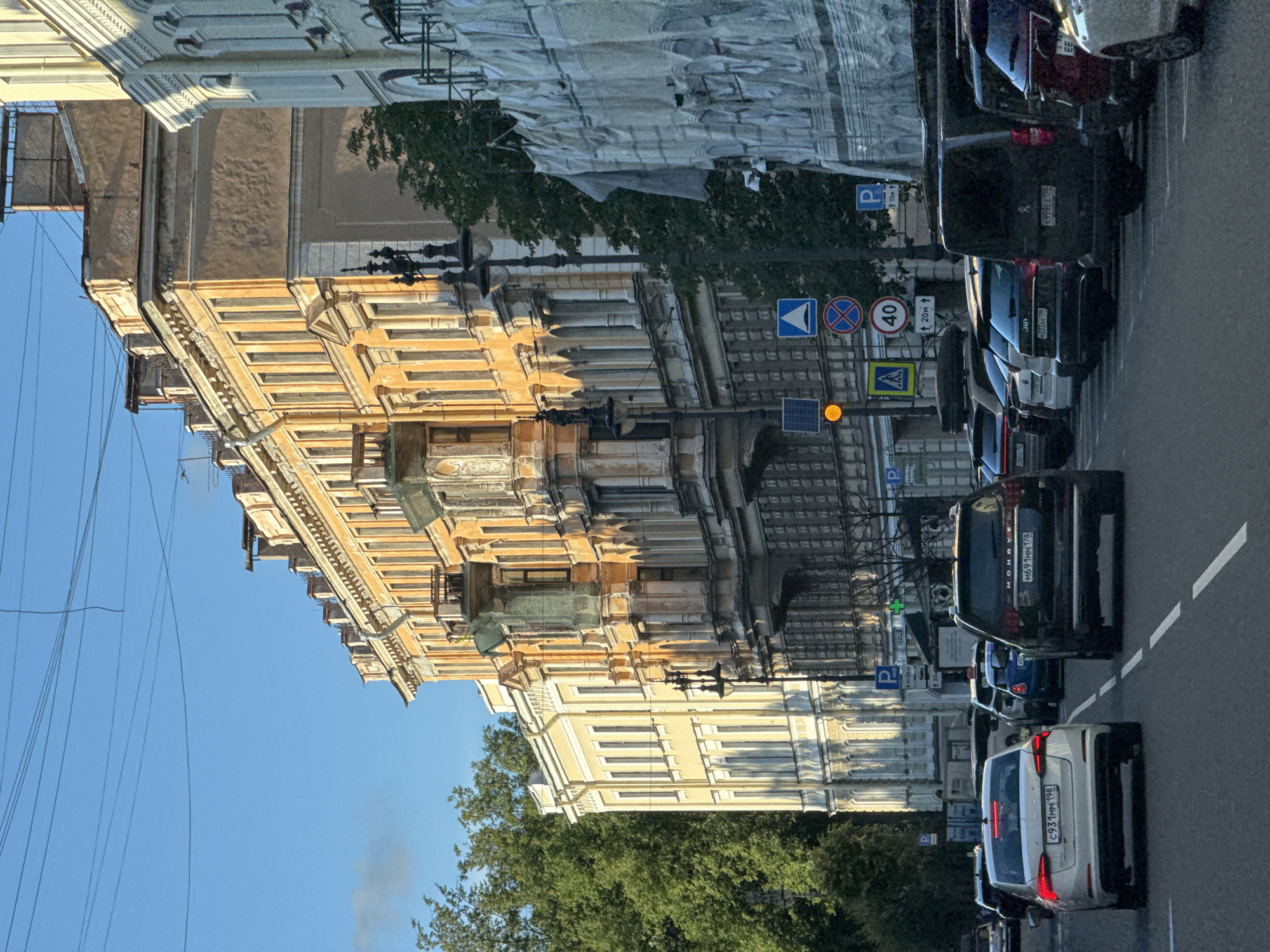 The image depicts a street scene in Saint Petersburg, Russia, likely during daylight hours with a clear blue sky. The architecture suggests a historic district, characterized by multi-story buildings with ornate facades, some of which are painted in yellow and ochre tones. One building on the right appears to be under renovation, covered in white sheeting. A line of parked cars, including a white sedan, a black SUV, and several other vehicles, is visible along the side of the street. Traffic signs, including speed limit signs (40 km/h) and directional indicators, are present. Trees and overhead electrical wires are also visible, contributing to the urban environment. The lighting indicates the sun is casting shadows, suggesting it is either morning or late afternoon.