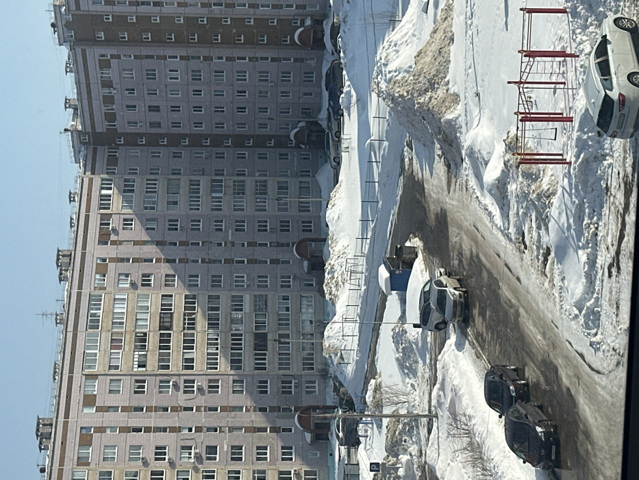 This image depicts a winter residential area in Chaikovsky, Russia, under a clear blue sky. Two large, multi-story apartment buildings, constructed with light-colored blocks or panels, form the background. They feature numerous white-framed windows and some enclosed balconies; the left building exhibits a curved architectural corner. The ground is extensively covered in snow, forming large, irregular piles, particularly evident in the mid-ground, suggesting snow removal operations. Asphalt roadways are partially clear, revealing wet and icy surfaces.
Multiple vehicles are present: a white Lada Vesta SW Cross station wagon is parked in the lower right, facing away from the viewer. On a winding road in the mid-ground, two dark sedans are parked on the left, followed by a silver minivan/SUV and a smaller white car behind it. Additional vehicles are parked in snow-covered areas in the background, near the apartment buildings.
A children's playground, comprising red metal climbing bars and a single swing, is located in the lower-mid ground, partially obscured by snow. A blue portable toilet with snow on its roof stands next to a trash bin in the mid-ground, adjacent to a cleared path. A "P" parking sign is visible on a pole to the left, partially obscured by snow. No individuals are observed within the scene.