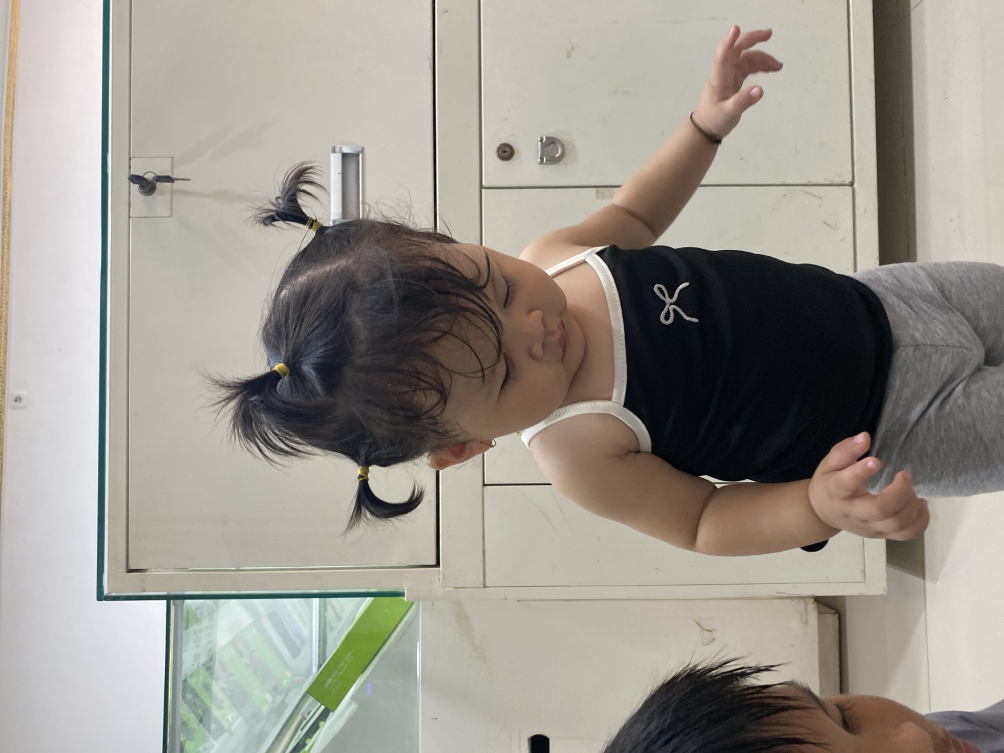 An Indonesian toddler with dark hair styled in pigtails is looking down and slightly to her right. She is wearing a black tank top with white trim and a white bow design on the front, paired with gray leggings. Her right arm is extended, and her hand is open. To her left, the head and shoulder of another person, possibly an older child, are partially visible. They have dark hair and are looking down. In the background is a beige metal filing cabinet with two drawers, each with a handle and a lock. A glass display case with green items inside is visible to the left of the filing cabinet. The flooring appears to be light-colored tile. The scene is indoors, likely in a home or office.