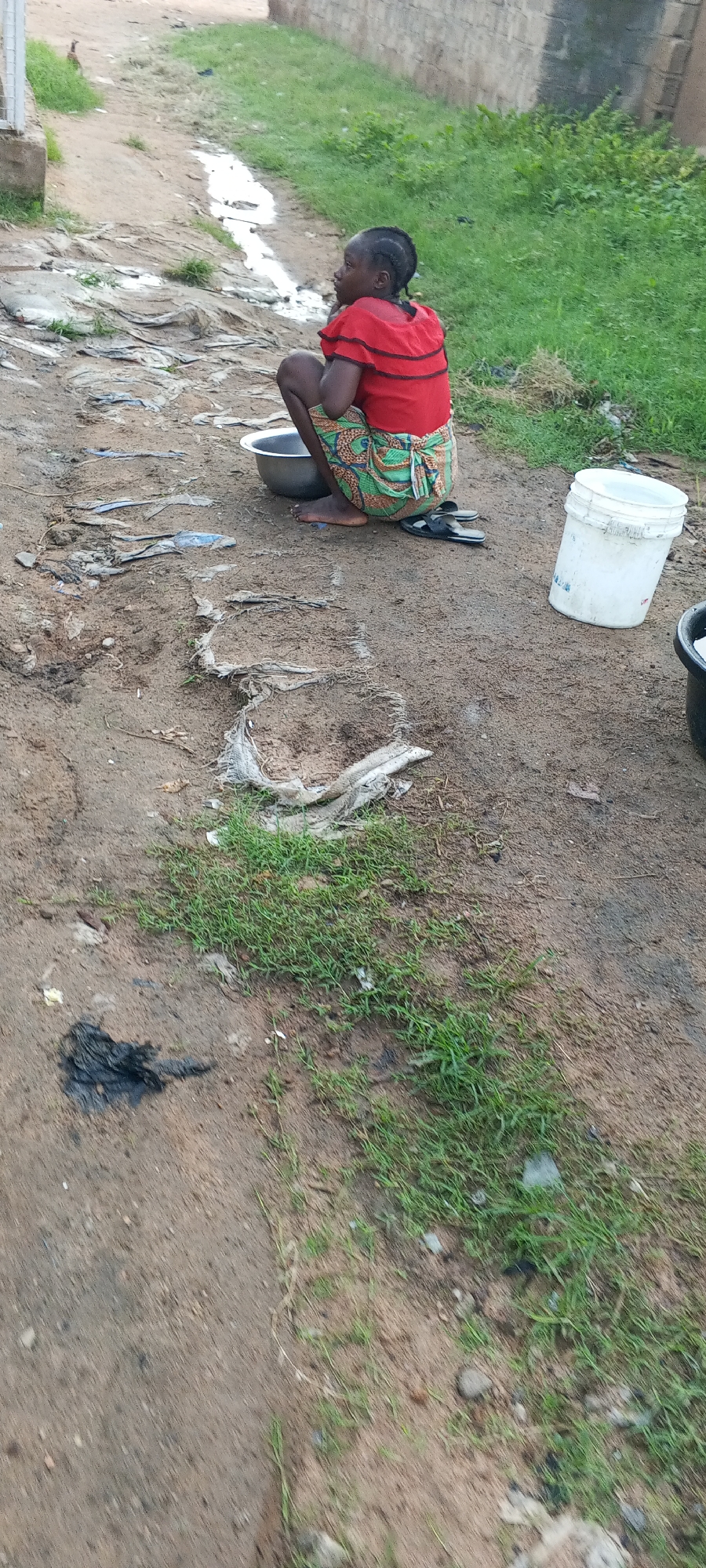 A young girl is squatting on the side of a dirt road in Jalingo, Nigeria. She is wearing a red shirt with black trim and a colorful patterned skirt. She has her hands resting on her knees and is looking to her left. In front of her is a silver basin, and next to her is a white bucket. The ground around her is dirt with patches of grass and some scattered debris, including what appears to be plastic bags. In the background, there is a low wall and more grass. The lighting suggests it is daytime.