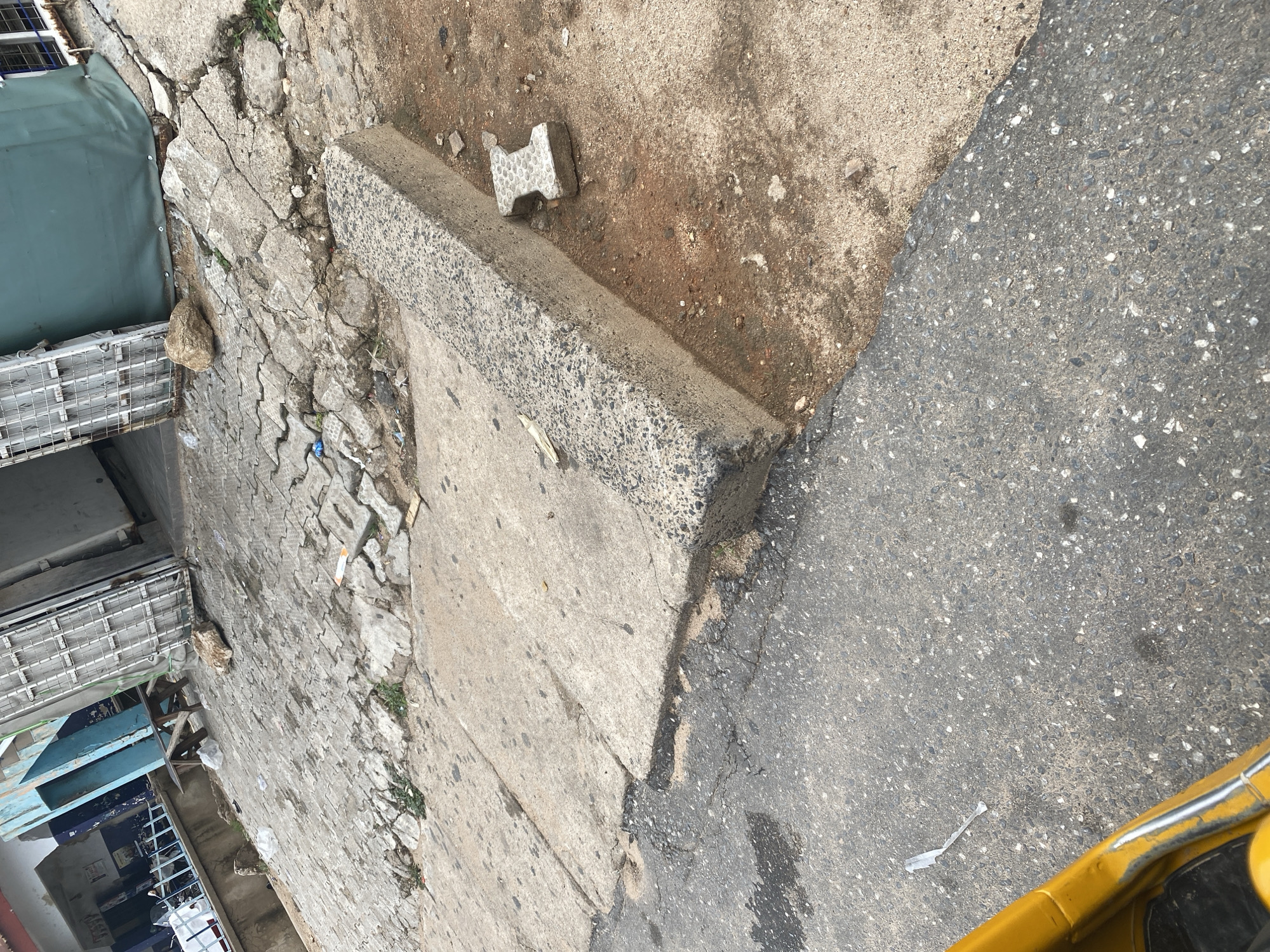 The media file shows an outdoor scene with a focus on a damaged concrete sidewalk and an adjacent asphalt road. The sidewalk is cracked and uneven, with some pieces broken away. A small, H-shaped interlocking paver lies on the ground next to a raised concrete curb. Beyond the sidewalk, there are glimpses of what appear to be market stalls or shops with metal shelving and blue and green coverings. A large rock is visible on the broken sidewalk. The ground is a mixture of dirt and concrete debris. The asphalt road is dark and appears wet in places, suggesting recent rain or a spill. In the lower right corner, a portion of a bright yellow vehicle is visible, possibly a tuk-tuk or a similar three-wheeled vehicle common in Nigeria. The scene appears to be located in an urban or semi-urban environment. The lighting suggests it is daytime, possibly with overcast conditions given the lack of harsh shadows. There is no discernible text in the image. The overall impression is one of a somewhat neglected urban street or market area.